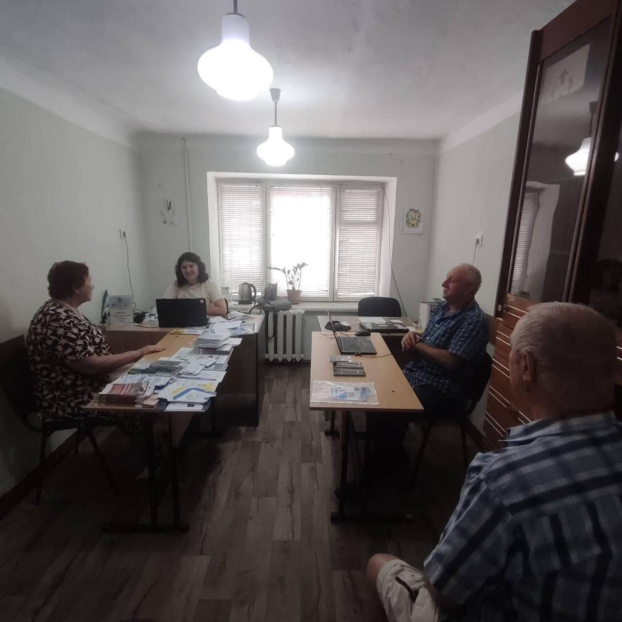 Начальниця ЦНАП Виноградівської сільської ради С.В. Сисоєва та адміністратор ЦНАП координаційного центру В.І. Чебан проводять прийом громадян у службовому кабінеті координаційного центру «Вільні разом» у місті Чернівці. У світлій кімнаті паралельно розташовані два дерев'яні столі. За лівим столом сидить С.В. Сисоєва за відкритим чорним ноутбуком, консультуючи літню відвідувачку в темній сукні з візерунком. На їхньому столі розкладено велику кількість паперових буклетів, листівок та бланків. За правим столом сидить В.І. Чебан у синій сорочці в клітинку, який також працює за ноутбуком і спілкується з чоловіком-переселенцем з Херсонщини. На передньому плані праворуч спиною до камери сидить ще один відвідувач у світлій сорочці в клітинку.