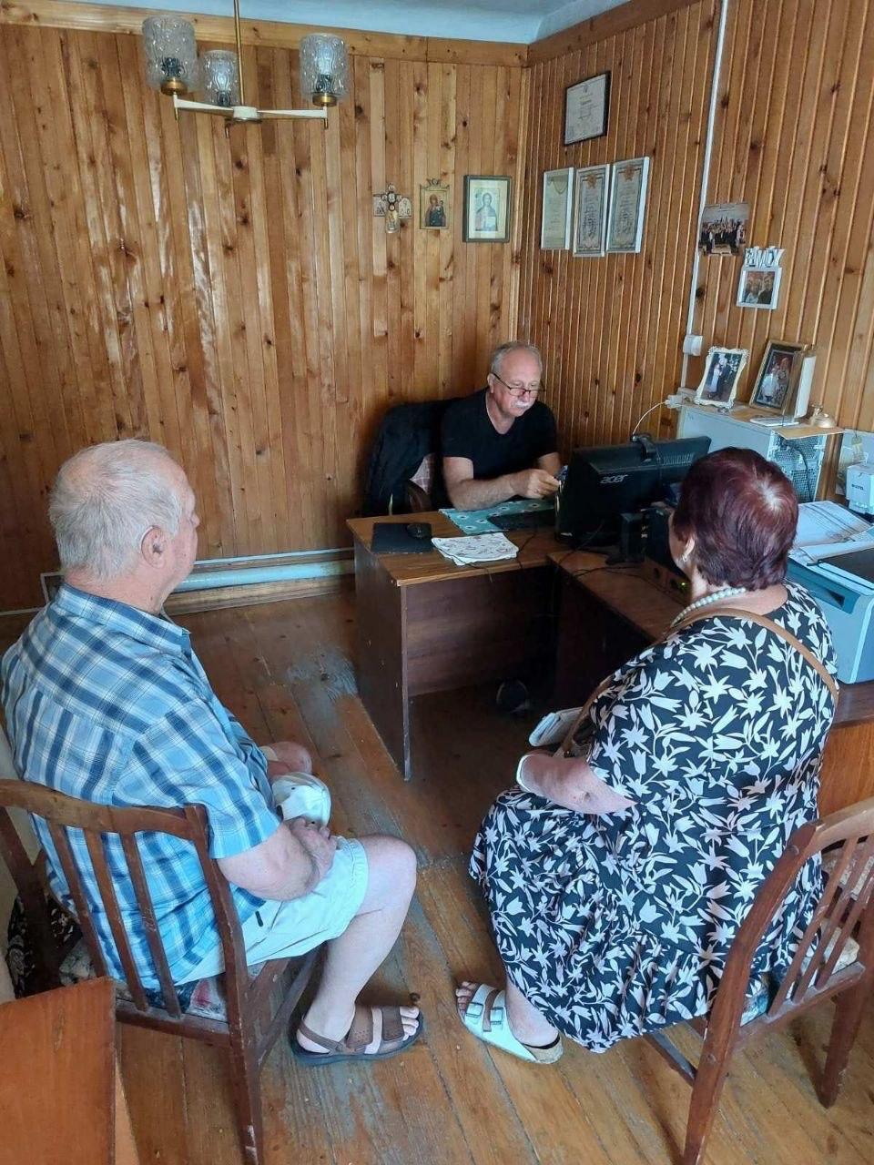 Юрист-волонтер проводить індивідуальний прийом та надає правові консультації парі літніх переселенців із Херсонщини в координаційному центрі «Вільні разом» у місті Чернівці. Зустріч проходить у робочому кабінеті, стіни, підлога та меблі якого повністю оздоблені світлим деревом (дерев'яною вагонкою). За великим кутовим столом сидить чоловік-юрист у чорній футболці та окулярах, який працює за комп'ютером бренду Acer. Навпроти нього на дерев'яних стільцях спиною до камери сидять двоє відвідувачів, які раніше отримували послуги ЦНАП: ліворуч — чоловік у синьо-білій сорочці в клітинку, праворуч — жінка в темній сукні з білим рослинним візерунком та з намистом на шиї.