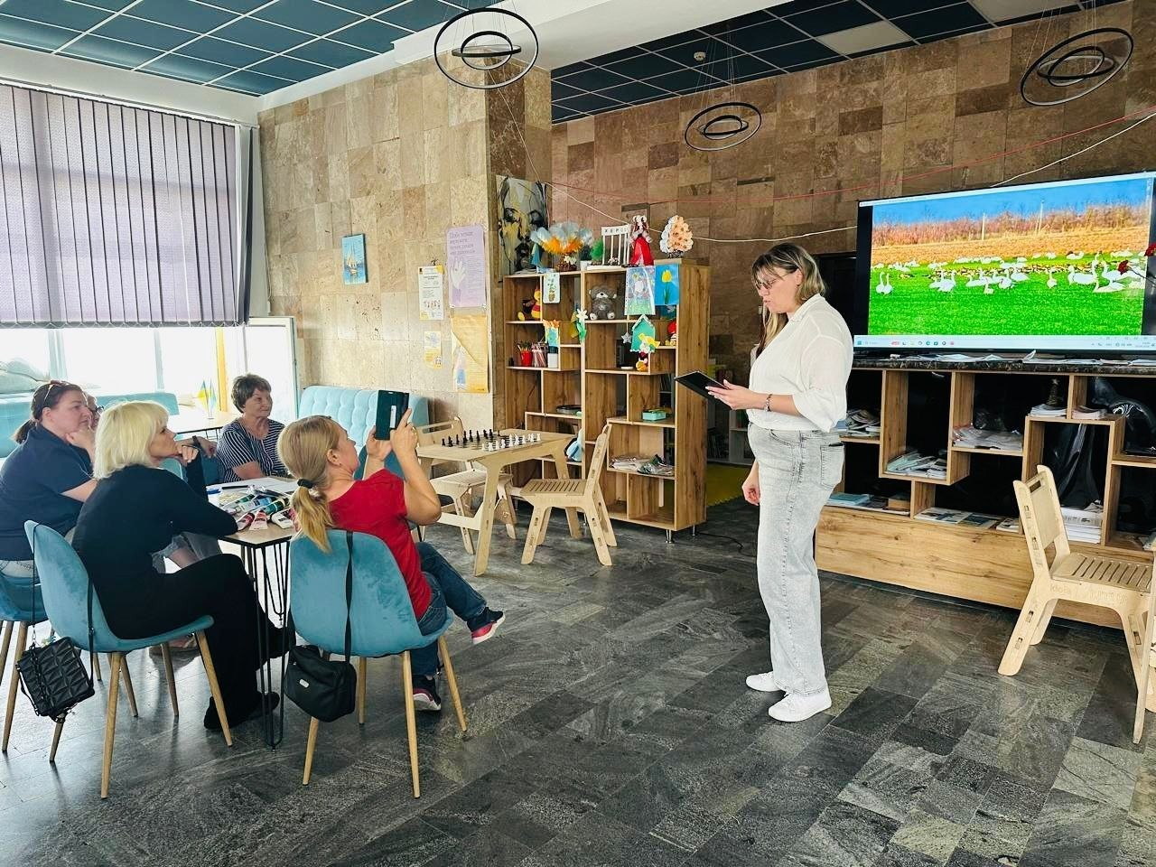 Презентація документальної аудіальної виставки «Справжній степ» для переселенців із Херсонщини в залі миколаївського координаційного центру «Вільні разом». Праворуч стоїть організаторка в окулярах, білій сорочці та світлих джинсах, яка тримає в руках чорний планшет і веде розповідь. Поруч із нею встановлено великий мультимедійний екран, на якому відображено яскравий краєвид зі зграєю білих птахів на зеленому полі, що символізує природу таврійського степу. Ліворуч за столом на м'яких бірюзових стільцях сидять чотири слухачки, які занурюються в атмосферу біосферного заповідника «Асканія-Нова» через аудіозаписи степових звуків. Одна з учасниць у червоній футболці знімає захід на свій смартфон.