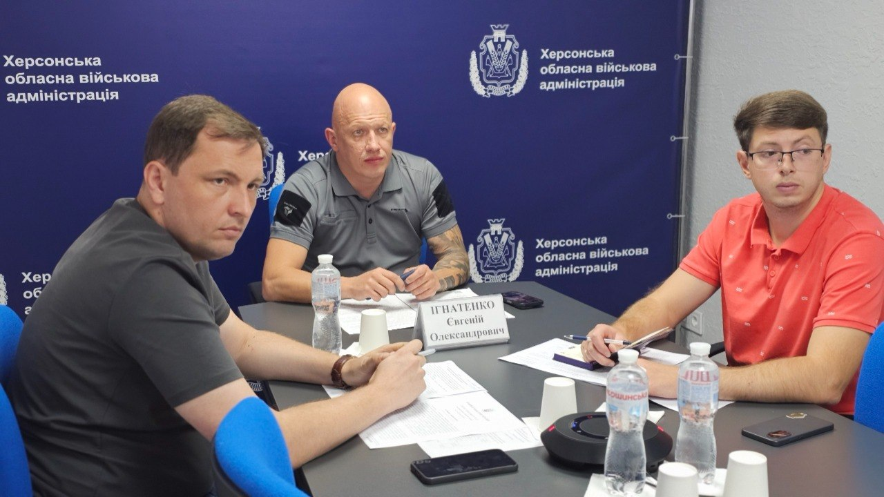 Заступник начальника Херсонської обласної військової адміністрації Євгеній Ігнатенко, директор Департаменту з питань цивільного захисту та оборонної роботи ХОВА Євген Пісоцький та представник ОДА сидять за великим сірим столом під час урядової міжвідомчої онлайн-наради. Зустріч присвячена фінансуванню невідкладних заходів зі зміцнення обороноздатності держави й антидронового захисту доріг. На передньому плані ліворуч у профіль сидить Євген Пісоцький у чорній футболці, уважно слухаючи колег. По центру на задньому плані сидить заступник начальника ОВА Євгеній Ігнатенко у сірій сорочці-поло з нашивками на рукавах. Праворуч сидить молодий спеціаліст в окулярах та яскраво-червоній сорочці-поло, який тримає в руці ручку над робочими документами. Нарада проходить у кабінеті на тлі офіційного темно-синього банера з написами «Херсонська обласна військова адміністрація».