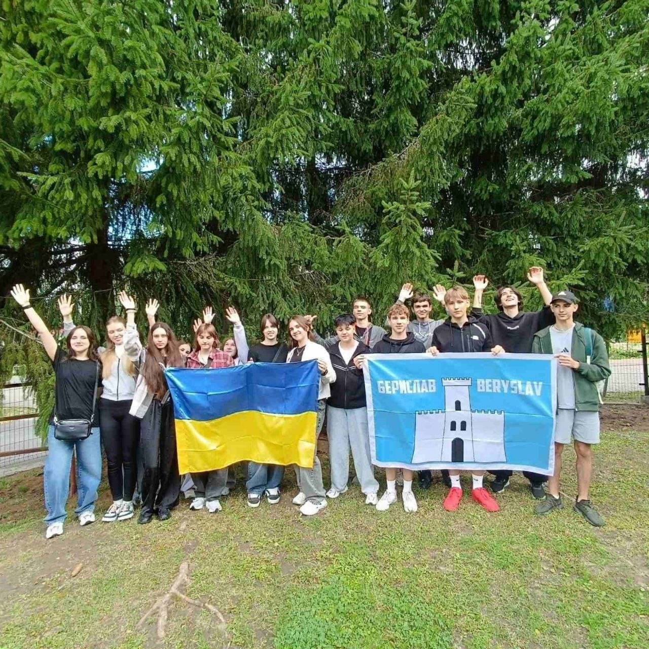 Група учнів Бериславського академічного ліцею позує для спільного патріотичного фото на відкритому повітрі під час візиту до Польщі в межах проєкту за підтримки Польсько-Української ради обміну молоддю. Підлітки стоять на трав'янистому майданчику на тлі великих густих зелених ялин, радісно посміхаються та підняли вгору руки у вітальному жесті. Попереду ліцеїсти розгорнули два прапори: ліворуч група дівчат тримає синьо-жовтий державний прапор України, а праворуч група хлопців — офіційний блакитний прапор міста Берислав із зображенням білої фортечної вежі в центрі та написами «БЕРИСЛАВ» і «BERYSLAV». Школярі прифронтової громади Херсонщини одягнені у повсякденний літній та спортивний одяг (джинси, футболки, худі, шорти).