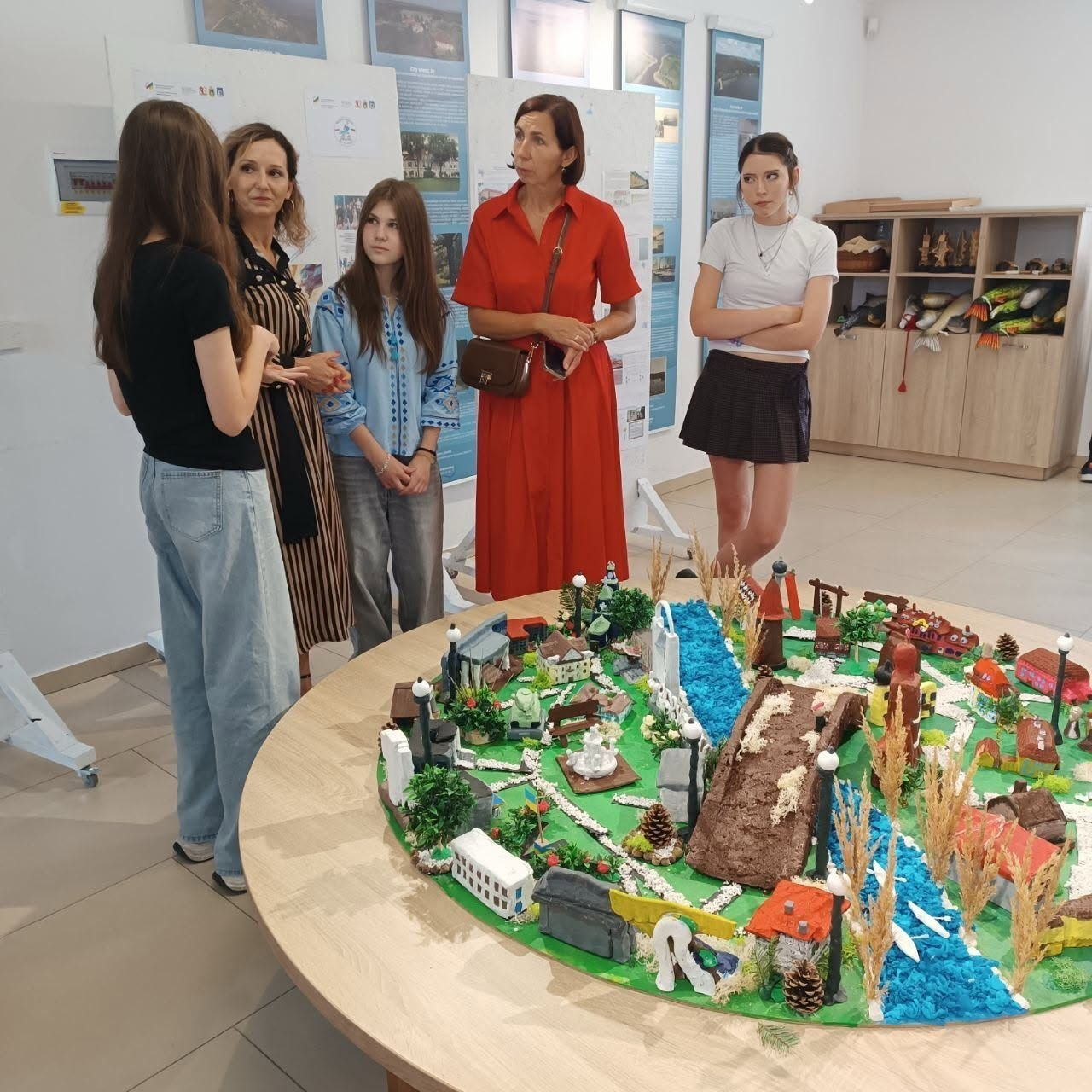 Презентація готового спільного проєкту «Місто Надії» у виставковій залі Музею народної культури у Венгожеві. Група з п'яти дівчат та жінок — представниць Бериславського академічного ліцею та польських партнерів — стоять навколо великого круглого дерев'яного столу й обговорюють результати роботи. На столі розгорнуто великий деталізований макет мініатюрного міста, створений ліцеїстами з глини, паперу, колосків пшениці та штучної зелені, де поміж мініатюрних кольорових будинків Берислава та Венгожева протікає річка з синього матеріалу. Зліва на передньому плані спиною до камери стоїть дівчина у чорній футболці та світлих широких джинсах. Поруч із нею стоять жінка у сукні у вертикальну смужку, дівчина у світло-блакитній вишиванці, жінка у довгій яскраво-червоній сукні та дівчина у білій футболці й темній короткій спідниці, яка схрестила руки на грудях. На задньому плані вздовж білих стін зали розміщено інформаційні стенди з фотографіями та картами.