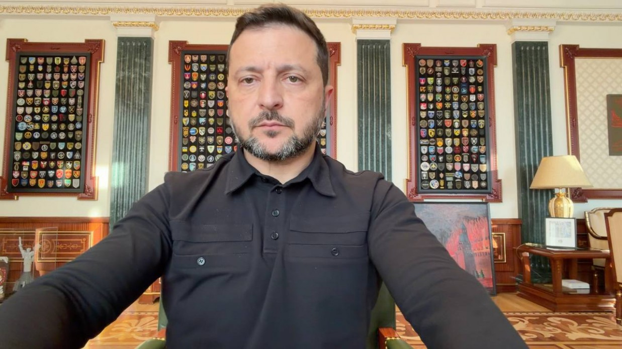 Президент України Володимир Зеленський зафіксований крупним планом по пояс під час щоденного офіційного відеозвернення до нації. У своїй промові глава держави повідомляє, що спецпосланці Президента США Дональда Трампа Стів Віткофф та Джаред Кушнер найближчими днями відвідають Москву, а в неділю прибудуть до Києва для пошуку реальних дипломатичних варіантів завершення війни. Володимир Зеленський одягнений у чорну тактичну сорочку-поло з довгими рукавами й нагрудними кишенями та сидить у кріслі свого робочого кабінету в Офісі Президента.