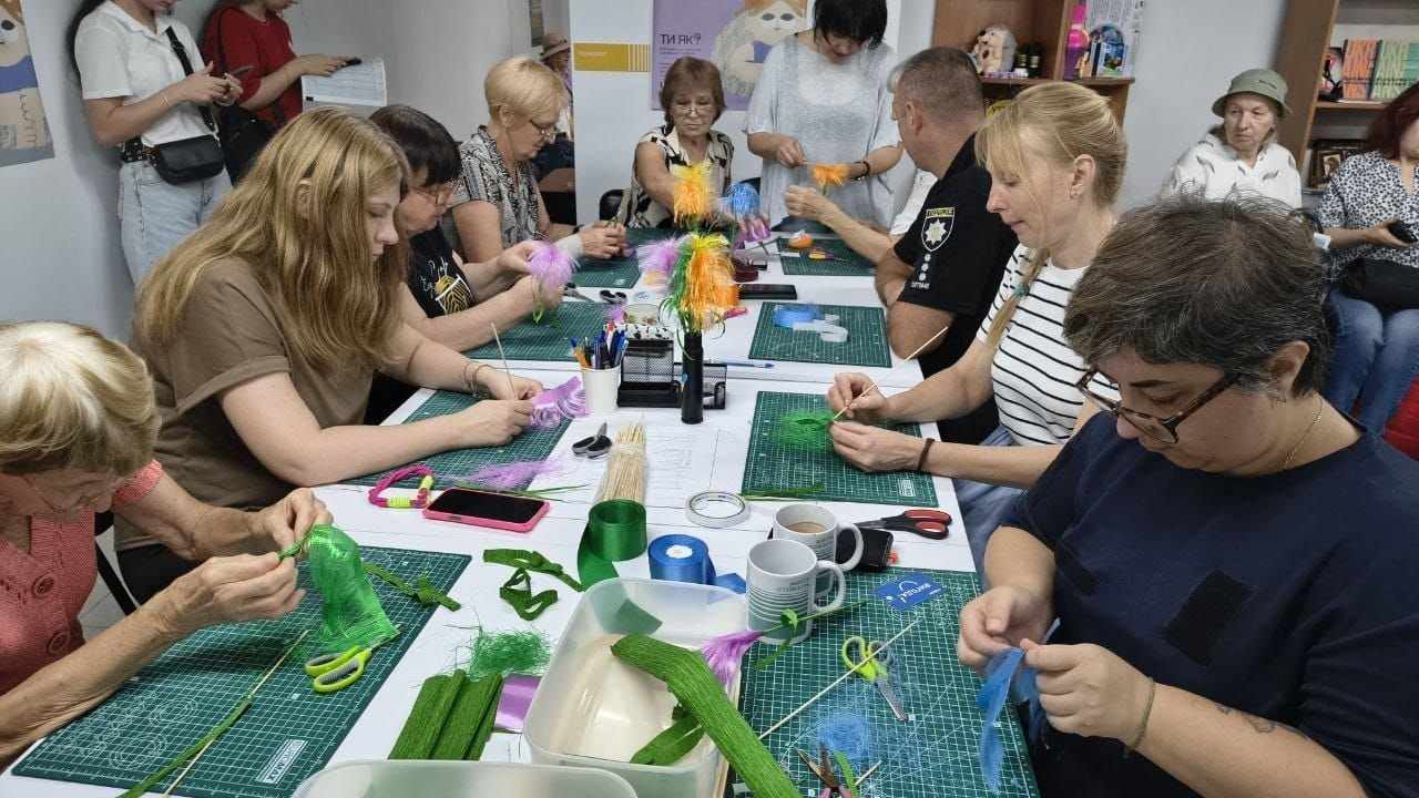Учасники різного віку та поліцейський під час творчого майстер-класу зі створення оберега «Домовик» у межах арттерапевтичних занять у Центрі життєстійкості. За великим білим столом з обох боків сидять жінки та підлітки, які зосереджено виготовляють саморобки з яскраво-зеленого гофрованого паперу, фіолетових і помаранчевих ниток та синіх стрічок. По центру праворуч за столом сидить чоловік у чорній формі Національної поліції України з офіційним шевроном на рукаві, який також бере участь у творчому процесі. На столі перед кожним учасником розкладено спеціальні зелені килимки для моделювання, ножиці, клей та дерев'яні шпажки. На білій стіні на задньому плані висить світлий патріотичний плакат Всеукраїнської програми ментального здоров'я з помітним написом «ТИ ЯК?».