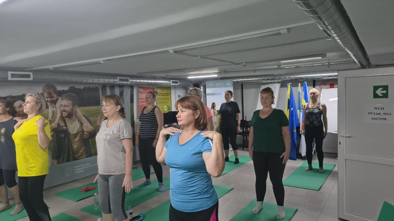 Група жінок різного віку під час оздоровчого заняття з пілатесу у спеціально облаштованому залі Центру життєстійкості. Учасниці тренування стоять у кілька рядів на яскраво-зелених спортивних килимках (каріматах) у просторому приміщенні з низькою стелею. На передньому плані по центру стоїть жінка у блакитній футболці та чорних лосинах, яка виконує розминку, поклавши пальці рук на плечі. Ліворуч від неї — жінка в яскраво-жовтій футболці та жінка в сірій спортивній кофті, які синхронно повторюють рухи. Усі присутні одягнені у зручний літній спортивний одяг та шкарпетки. На задньому плані праворуч, біля білої маркерної дошки, встановлено державні прапори.