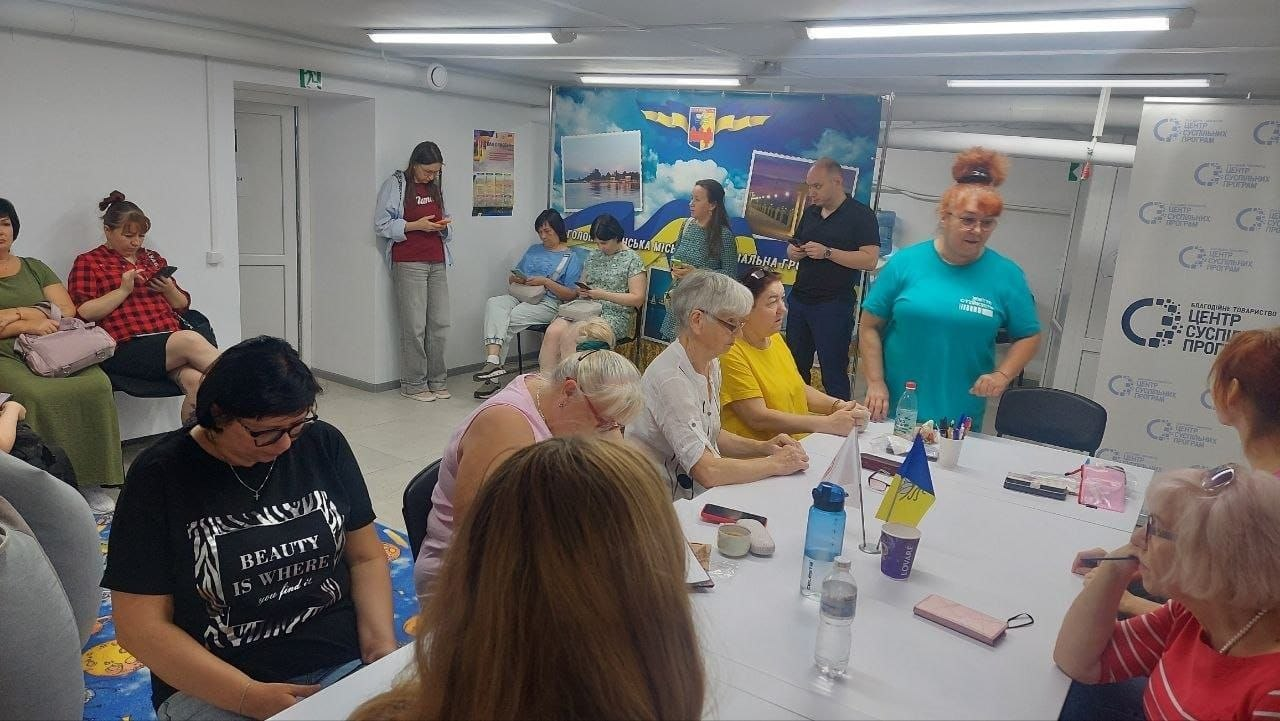 Психологічний захід у Центрі життєстійкості, присвячений розбору впливу стресу та техніці «Світлофор мого стану». У центрі кімнати за великим Т-подібним білим столом сидять жінки різного віку, які зосереджено слухають та роблять нотатки. Перед ними на столі розкладено блокноти, ручки, смартфони, стоять пляшки з водою та маленький настільний прапорець України. Праворуч стоїть жінка-тренерка в яскраво-бірюзовій футболці, яка веде бесіду з присутніми та роз'яснює вправи для «заземлення» й боротьби з тривогою. Уздовж стін на задньому та лівому планах на стільцях сидять інші учасниці зустрічі.