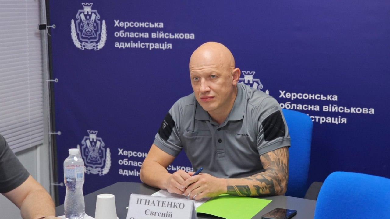 Заступник начальника Херсонської ОВА Євгеній Ігнатенко сидить за сірим робочим столом під час онлайн-наради з Міністерством економіки та довкілля України. Зустріч присвячена обговоренню проєкту урядового розпорядження про виділення 50 мільйонів гривень із резервного фонду держбюджету на фінансування інженерного захисту об’єктів критичної інфраструктури Херсонщини. Посадовець одягнений у сіру сорочку-поло з чорними елементами на плечах, його руки складені на столі, а в пальцях він тримає синю кулькову ручку. Перед ним лежить відкрита папка з яскраво-зеленими робочими аркушами, смартфон та встановлено білу іменну табличку. Засідання проходить у кабінеті на тлі офіційного темно-синього банера з повторюваними емблемами та написом Херсонська обласна військова адміністрація.