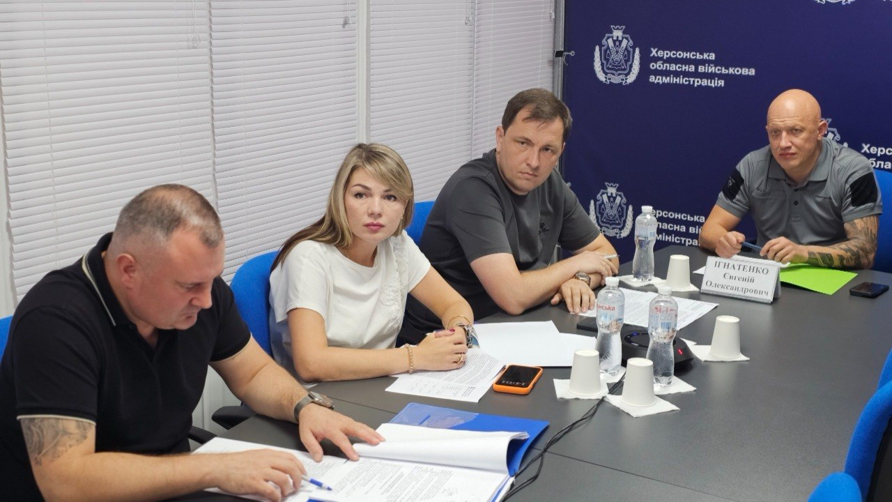 Робоча нарада щодо фінансування інженерного захисту критичної інфраструктури Херсонщини за участі представників ОВА. Чотири посадовці сидять за круглим столом у залі засідань на тлі синього брендованого банера з гербом та написами «Херсонська обласна військова адміністрація». Справа сидить заступник начальника Херсонської ОВА Євгеній Ігнатенко, одягнений у світло-сіру спортивну футболку-поло з темними вставками на плечах і комірі. Безпосередньо ліворуч від нього сидить директор Департаменту з питань цивільного захисту та оборонної роботи ХОВА Євген Пісоцький у темно-сірій звичайній футболці з невеликим принтом. Поруч із ними за столом перебувають представники профільних структурних підрозділів ХОДА: жінка зі світлим волоссям у класичній білій футболці та чоловік на передньому плані ліворуч, одягнений у чорну футболку-поло з білим кантиком на комірі, який вивчає роздруковані документи проєкту урядового розпорядження про спрямування 50 мільйонів гривень на захист об’єктів.