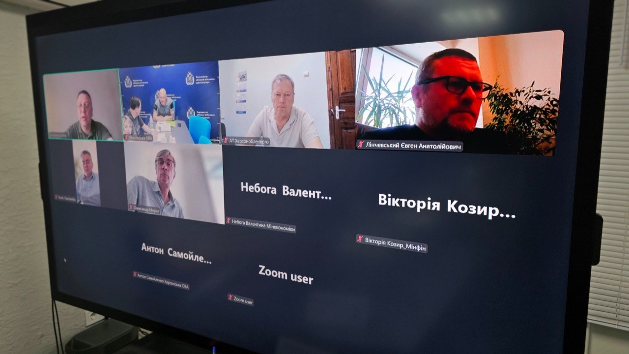 Екран монітора з онлайн-нарадою у програмі Zoom щодо виділення 50 мільйонів гривень на інженерний захист критичної інфраструктури Херсонщини. На великому дисплеї відображається сітка з вікнами учасників зустрічі. У верхньому ряду ліворуч транслюється відео із зали засідань Херсонської ОВА за участі заступника начальника ОВА Євгенія Ігнатенка та директора Департаменту цивільного захисту Євгена Пісоцького, а також вікно представника АТ «Херсонобленерго». Решта вікон відображають текстові підписи представників профільних відомств без увімкнених камер, серед яких представниця Мінекономіки Валентина Небога, представниця Мінфіну Вікторія Козир та ще один заступник начальника Херсонської ОВА Антон Самойленко. Учасники онлайн-формату погоджують проєкт урядового розпорядження для виділення коштів із резервного фонду держбюджету.