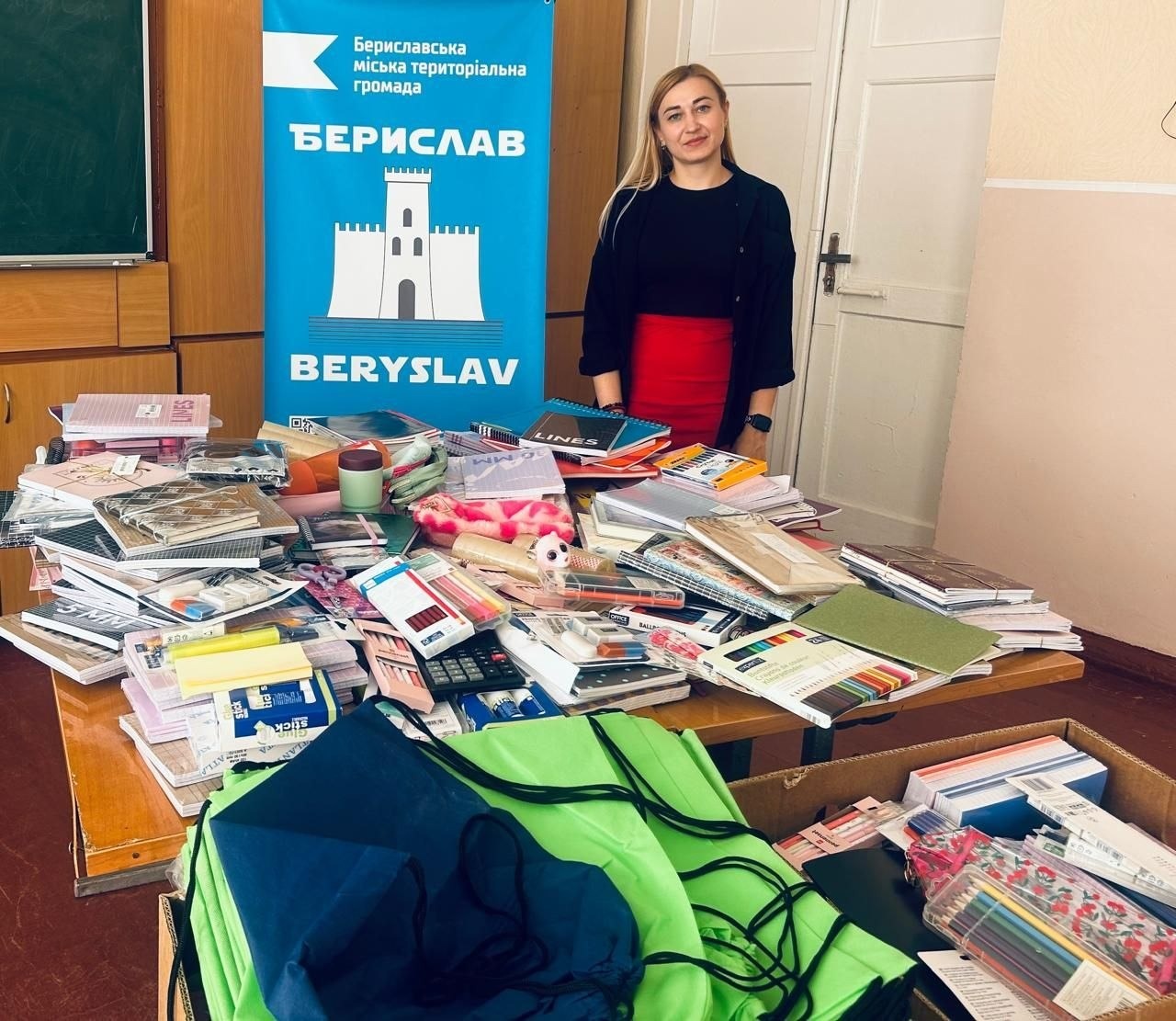 Канцелярське приладдя та шкільні набори для школярів Бериславської громади, отримані як благодійна допомога від партнерів із Нідерландів. На фотографії зображена представниця громади у чорній кофті та червоній спідниці, яка стоїть у кімнаті біля великого столу на тлі синього вертикального банера з гербом міста та написом «Бериславська міська територіальна громада. БЕРИСЛАВ. BERYSLAV». Весь стіл перед нею повністю заповнений канцелярським приладдям: зошитами, блокнотами, кольоровими олівцями, ручками, калькуляторами та клеєм. На передньому плані на підлозі стоїть коробка з яскраво-зеленими тканинними рюкзаками-мішками в межах ініціативи «Rugzakjes vol Hoop» («Рюкзачки, сповнені надії») для юних жителів Херсонщини, зібраними мешканцями та організаціями міста-партнера Вінтерсвейк.
