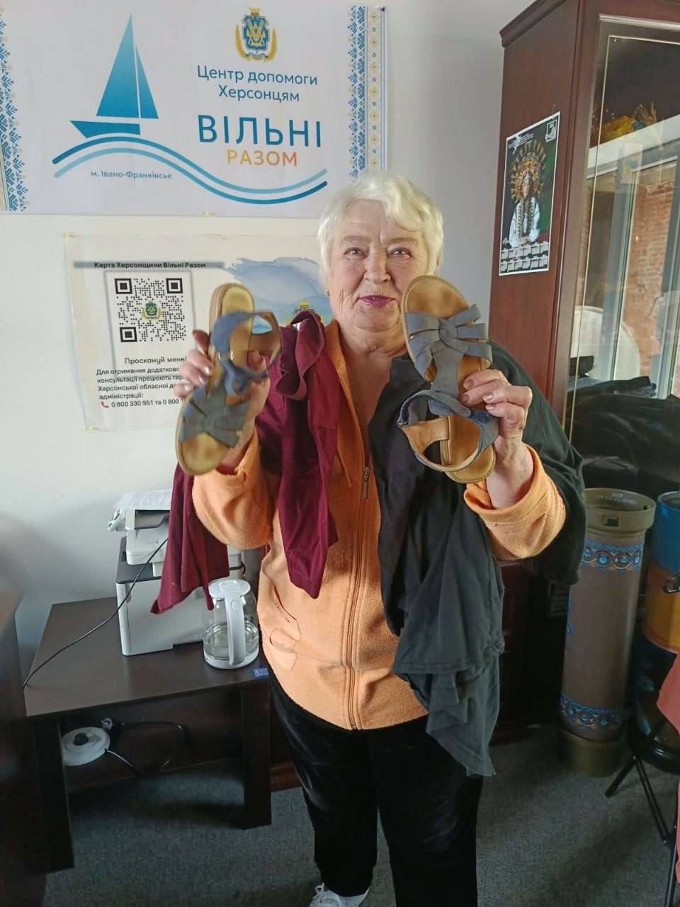 Літня переселенка з Херсонщини отримує гуманітарну допомогу від Фонду громади міста Херсон «Захист» у координаційному центрі «Вільні разом» в Івано-Франківську. Жінка зі світлим коротким волоссям, одягнена в персикову кофту на блискавці та чорні штани, стоїть посеред кімнати і тримає в обох руках отриманий вживаний сезонний одяг та пару синіх жіночих літніх босоніжок на плоскій підошві. Позаду неї на стіні розміщено офіційний плакат Центру допомоги херсонцям «ВІЛЬНІ РАЗОМ» із зазначенням міста Івано-Франківськ, а нижче висить інформаційний листок «Карта Херсонщини Вільні Разом» із QR-кодом для отримання консультацій.