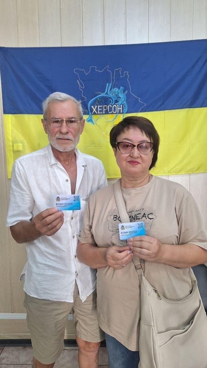 Двоє внутрішньо переміщених осіб старшого віку з Херсонщини позують для фото під час реалізації проєкту Картка Херсонщини в одеському координаційному центрі Вільні разом. Праворуч стоїть жінка в окулярах та світло-бежевій футболці з великою сумкою через плече, яка з посмішкою тримає перед собою блакитну пластикову Картку Херсонщини. Ліворуч стоїть сивочолий чоловік в окулярах, білій літній сорочці та шортах, який так само демонструє отриману картку з логотипом центру Вільні разом. Позаду них на стіні висить синьо-жовтий державний прапор України, у центрі якого нанесено контурні обриси карти Херсонської області, стилізовані під судини серця, та напис ХЕРСОН.