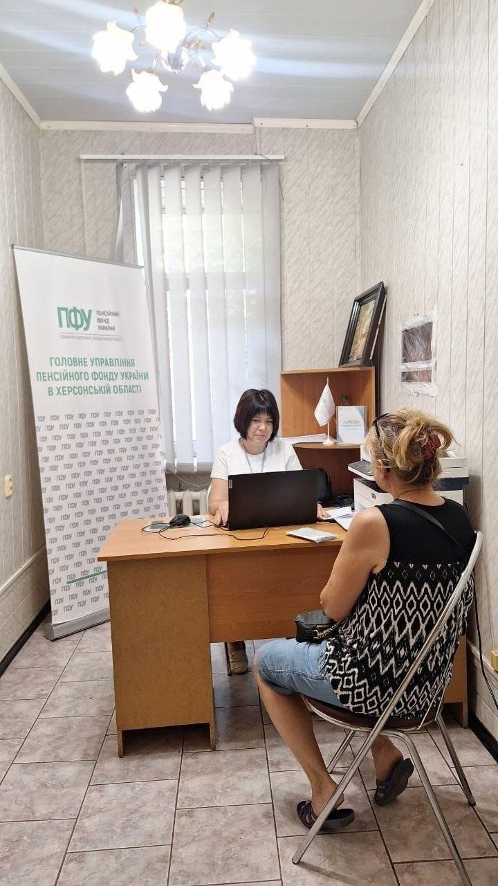 Виїзний прийом громадян фахівцем Пенсійного фонду України для внутрішньо переміщених осіб із Херсонщини в одеському координаційному центрі Вільні разом. За дерев'яним робочим столом сидить жінка-консультант у білій футболці, яка працює за відкритим чорним ноутбуком та надає роз'яснення відвідувачці. Навпроти неї спиною до камери на металевому стільці сидить жінка у темній майці з геометричним візерунком та джинсових шортах. Ліворуч від робочого столу встановлено великий мобільний ролап-банер Головного управління Пенсійного фонду України в Херсонській області з фірмовим зеленим логотипом ПФУ. Вікно на задньому плані закрите світлими вертикальними жалюзі, а на поличці праворуч розміщено документи, картину в рамці та маленький білий прапорець на підставці.
