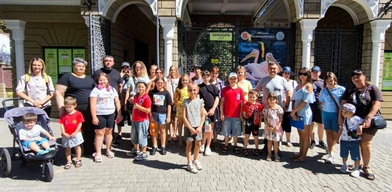 Велика група родин із дітьми — внутрішньо переміщених осіб із Херсонщини — позують для спільного фото під час безкоштовного екскурсійного візиту до Одеського зоопарку. Захід організовано херсонським координаційним центром Вільні разом в Одесі. Діти, підлітки та дорослі супроводжуючі стоять у кілька рядів на брукованій площі перед головним входом у сонячний літній день. Усі учасники одягнені у повсякденний літній одяг (футболки, шорти, сукні та кепки). З лівого краю на передньому плані посміхається маленький хлопчик, який сидить у триколісному дитячому візку. На задньому плані чітко видно масивні ковані ворота й кам'яні арки входу до зоопарку, де встановлено зелену вивіску ЗООПАРК та великий круглий банер із зображенням рожевих пеліканів та ювілейним написом.