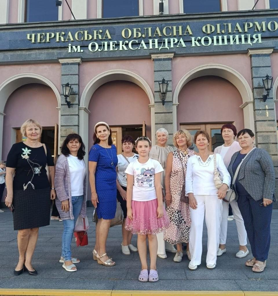Група з дев'яти жінок та однієї дівчинки позує для спільного фото на тлі парадного входу до Черкаської обласної філармонії імені Олександра Кошиця. Учасниці зустрічі стоять у ряд на брукованому майданчику в сонячний день, одягнені у святковий та повсякденний літній одяг. По центру на передньому плані стоїть дівчинка у білій футболці з малюнком та пишній рожевій спідниці з блискітками. Ліворуч від неї стоїть жінка у яскраво-синій літній сукні та світлому обручі на голові. Інші жінки одягнені у сукні з принтами, світлі штани, блузи та кардигани. Будівля філармонії на задньому плані має рожевий фасад, великі арочні ніші з дерев'яними вхідними дверима та класичні вуличні ліхтарі на стінах. Над арками великими золотими літерами нанесено напис ЧЕРКАСЬКА ОБЛАСНА ФІЛАРМОНІЯ ім. ОЛЕКСАНДРА КОШИЦЯ.