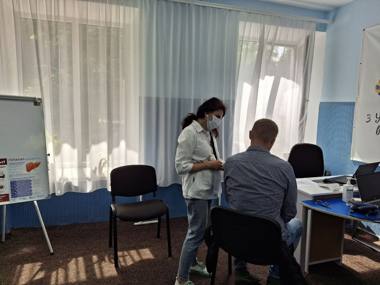 Медична консультація для внутрішньо переміщених осіб із Херсонщини в хмельницькому координаційному центрі Вільні разом, яку проводять лікарі комунального некомерційного підприємства Центр первинної медико-санітарної допомоги Рубіжанської міської ради. По центру стоїть жінка-лікар у білому медичному халаті, захисній масці та блакитних джинсах, яка спілкується з пацієнтом і проводить огляд. Чоловік сидить спиною до камери на чорному офісному стільці, він одягнений у синю сорочку в дрібну клітинку та джинси. Праворуч розташовано робочий стіл фахівця, де розкладено документи, стоїть відкритий ноутбук, тонометр для вимірювання артеріального тиску та флакон з антисептиком. На задньому плані кадру видно великі вікна зі світлою тюлю, а ліворуч встановлено мобільну дошку-фліпчарт з медичним інформаційним плакатом, присвяченим профілактиці та діагностиці гепатиту.