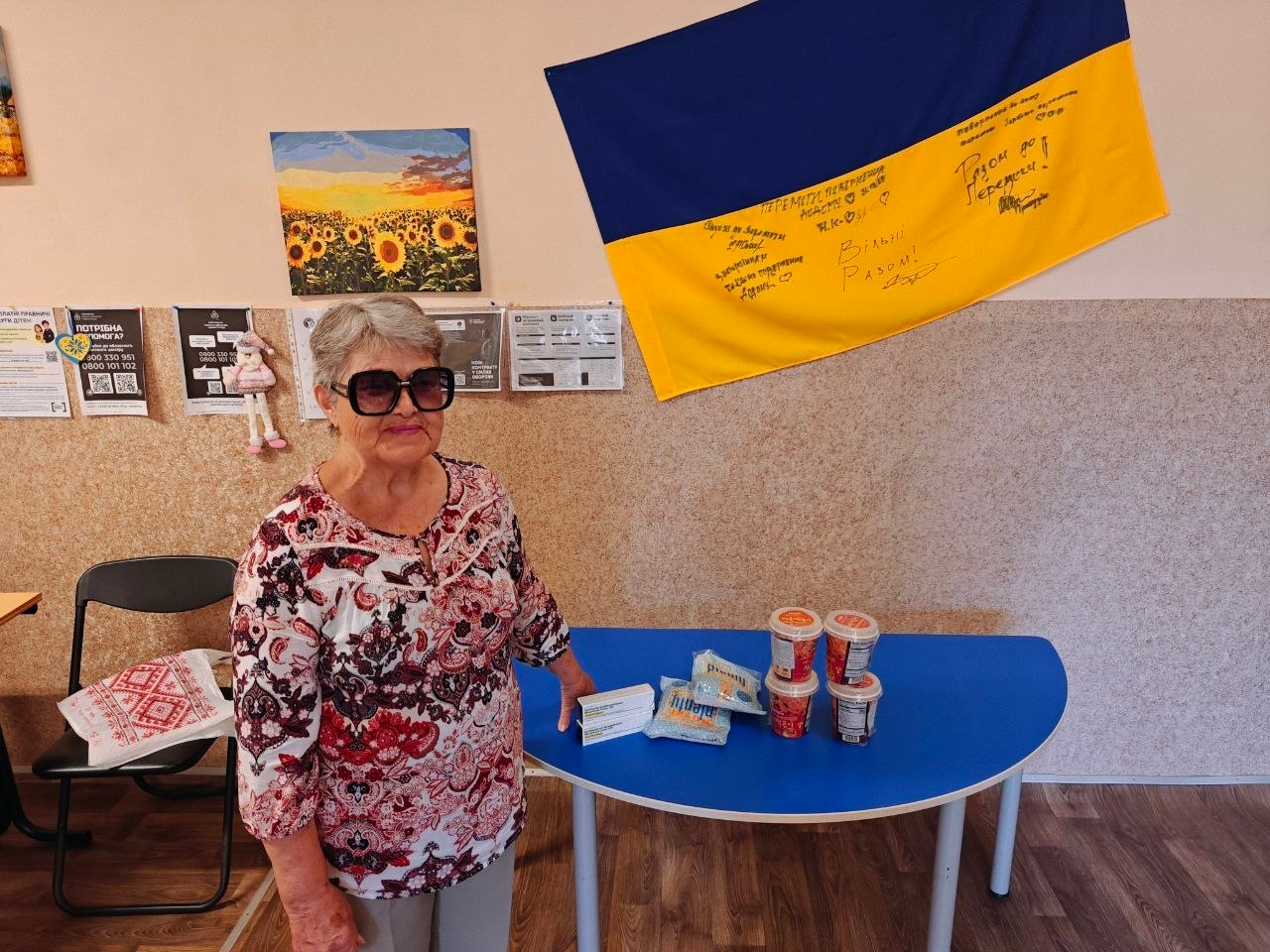 Літня жінка — внутрішньо переміщена особа з Херсонщини — отримує безкоштовні медикаменти після медичного огляду мобільною бригадою Товариства Червоного Хреста України в хмельницькому координаційному центру Вільні разом. Вона стоїть ліворуч у великих сонцезахисних окулярах і світлій блузі з біло-червоним квітковим візерунком. На синій поверхні напівкруглого столу перед нею розкладено отримані за результатами скринінгу лікарські засоби, пачка вологих серветок, а також баночки зі спеціалізованим медичним або дитячим харчуванням. На бежевій стіні позаду жінки висить великий державний прапор України з рукописними побажаннями від відвідувачів та волонтерів, серед яких слова Разом до Перемоги! та Вільні Разом!. Ліворуч на стіні розміщено невелику картину з соняшниковим полем на заході сонця та офіційні інформаційні плакати з контактами гарячих ліній допомоги для ВПО.