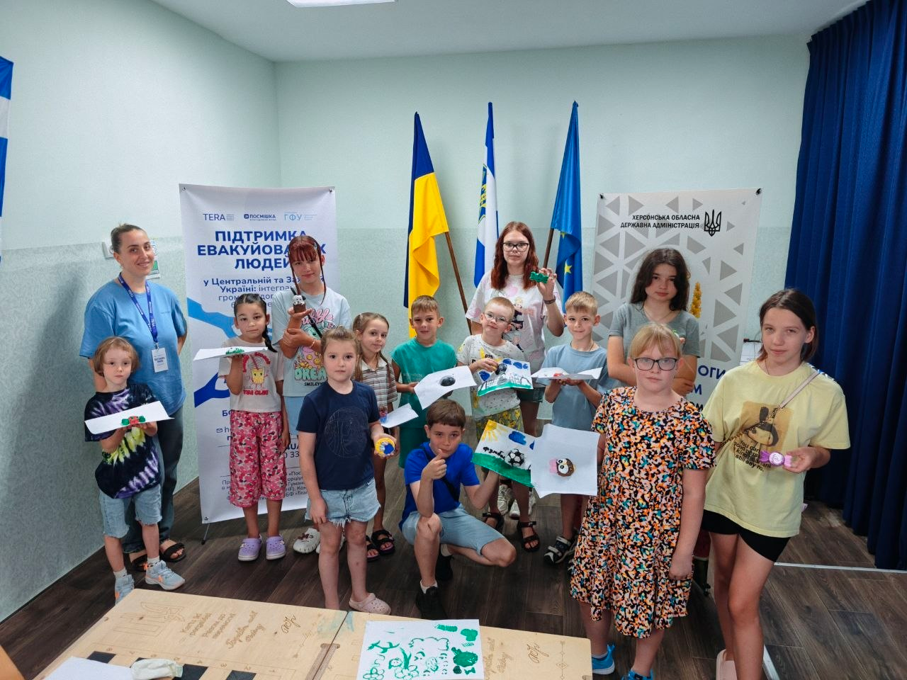 Учасники дитячого артзаняття У які кольори розфарбована мрія позують для спільного фото в хмельницькому координаційному центрі Вільні разом. Захід для тринадцяти дітей внутрішньо переміщених осіб із Херсонщини організовано за підтримки благодійного фонду Посмішка UA. Діти різного віку та дві жінки-координаторки стоять у просторій кімнаті, тримаючи в руках свої творчі роботи: малюнки на білих аркушах паперу та невеликі кольорові саморобки. На передньому плані по центру хлопчик у синій футболці присів навпочіпки, показуючи жест схвалення з піднятим догори великим пальцем. З лівого боку кадру стоїть представниця благодійної організації у блакитній футболці з офіційним бейджем на шиї. На задньому плані по центру приміщення встановлено три великі прапори на вертикальних флагштоках: державний прапор України, синьо-білий прапор Херсонської області та синій прапор Європейського Союзу. По обидва боки від прапорів розміщено два інформаційні ролап-банери: ліворуч — стенд фонду Посмішка UA з написом Підтримка евакуйованих людей, праворуч — офіційний білий банер із золотим тризубом та написом Херсонська обласна державна адміністрація. На передньому плані знизу видно краєчки робочих столів із розкладеними дитячими малюнками.