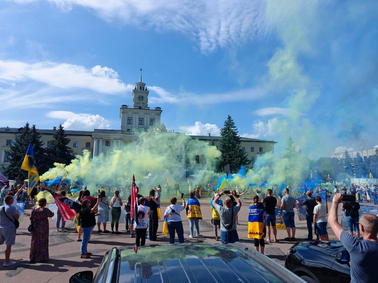 Учасники патріотичної акції та автопробігу на підтримку військовополонених, зниклих безвісти та їхніх сімей, організованого за участі хмельницького координаційного центру Вільні разом для внутрішньо переміщених осіб із Херсонщини. На великій міській площі перед будівлею Хмельницької обласної ради велика група людей стоїть спиною до камери, тримаючи державні прапори України, прапори військових підрозділів та патріотичні транспаранти й плакати. Уздовж лінії учасників у повітря вириваються густі клуби кольорового диму синього та жовтого кольорів, які створюють у небі масштабну імітацію українського прапора. На задньому плані височіє велична світла адміністративна будівля у класичному стилі з високою центральною вежею, шпилем та годинником, оточена високими зеленими ялинами. На передньому плані знизу частково видно дахи та капоти автомобілів учасників автопробігу під світло-блакитним сонячним небом.