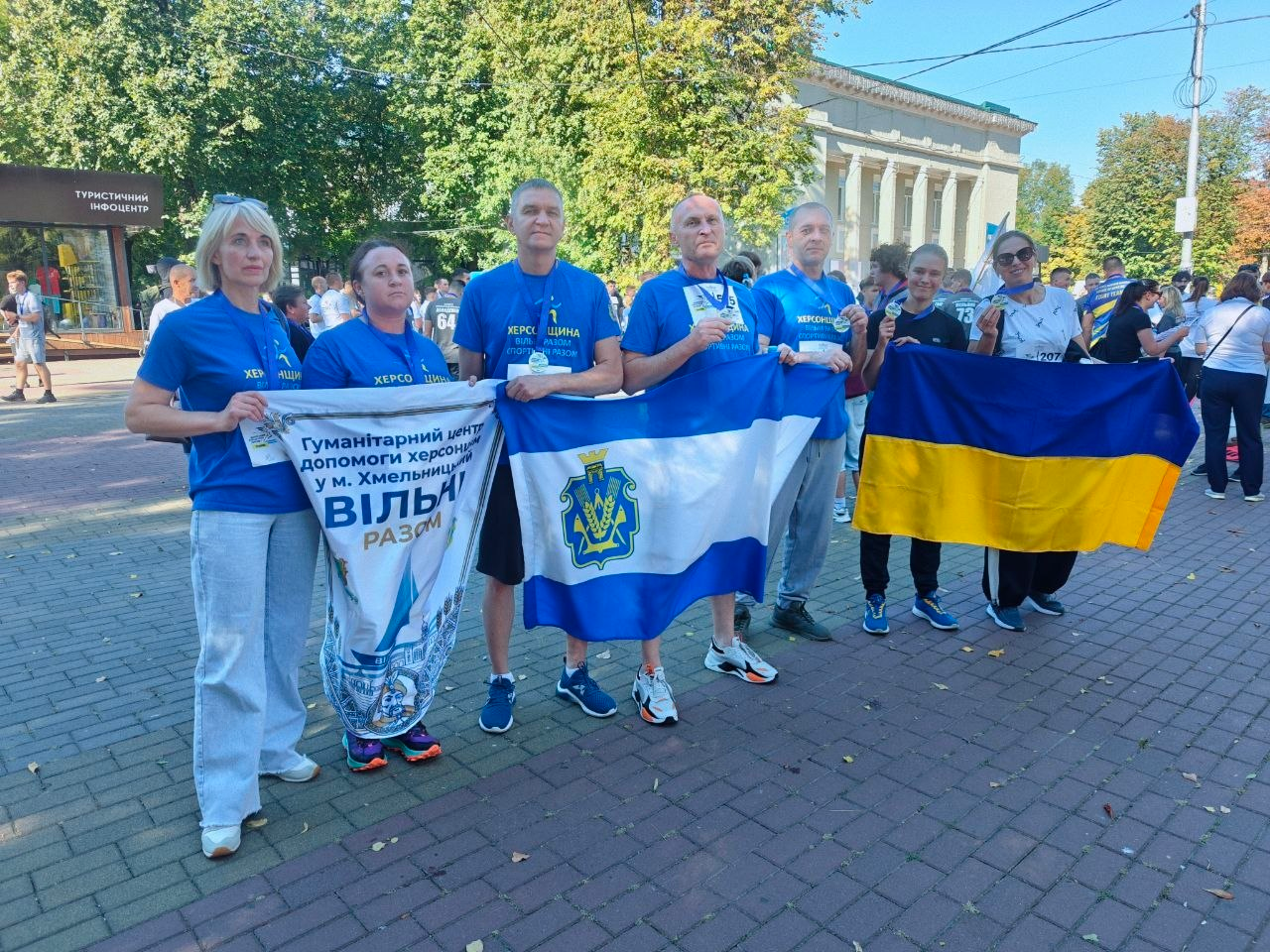 Представники та відвідувачі хмельницького координаційного центру Вільні разом — внутрішньо переміщені особи з Херсонщини — позують для спільного фото на брукованій міській алеї після участі в щорічному забігу Шаную воїнів, біжу за Героїв України, присвяченому Дню пам’яті захисників і захисниць України. Чоловіки та жінки стоять у ряд у синіх фірмових футболках із патріотичними логотипами та тримають у руках отримані медалі учасників забігу. Крайні ліворуч дві жінки тримають білий вертикальний банер із написом Гуманітарний центр допомоги херсонцям у м. Хмельницькому ВІЛЬНІ РАЗОМ. По центру троє чоловіків розгорнули великий біло-синій прапор Херсонської області з офіційним гербом. Праворуч ще дві жінки тримають розгорнутий синьо-жовтий державний прапор України. На задньому плані кадру в сонячний день видно густі зелені дерева, будівлю у класичному архітектурному стилі, міську вивіску Туристичний інфоцентр та інших учасників масового патріотичного марафону.
