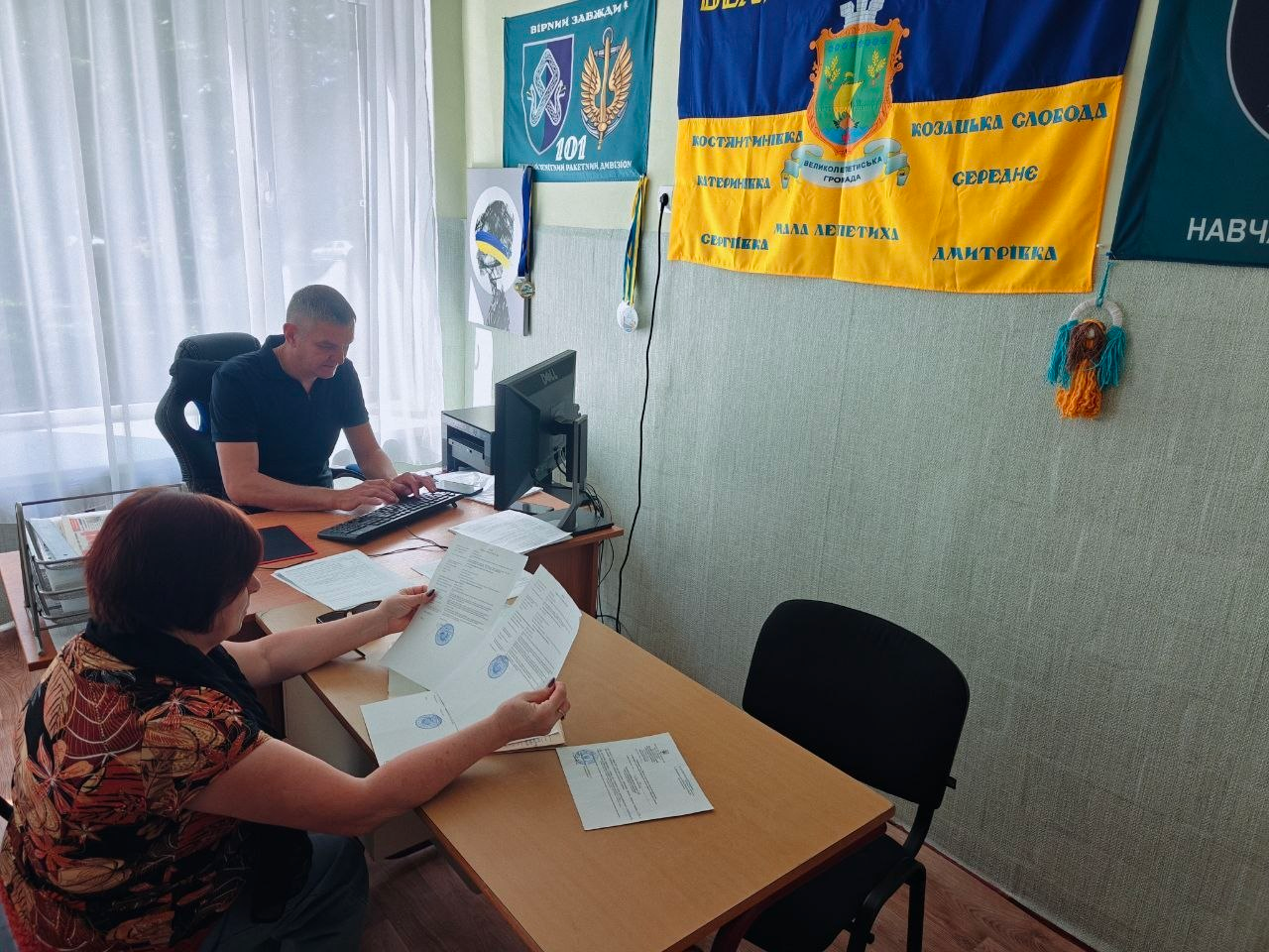 Прийом та консультування внутрішньо переміщених осіб із Херсонщини фахівцями Центру надання адміністративних послуг у хмельницькому координаційному центрі Вільні разом. За двома зсунутими дерев'яними столами в кабінеті сидять двоє людей. На задньому плані чоловік у чорній футболці-поло, працівник установи, опрацьовує звернення за комп'ютером, тримаючи руки на клавіатурі перед монітором Dell. На передньому плані ліворуч сидітиме жінка у різнокольоровій літній блузі, яка тримає в руках та уважно вивчає офіційні документи з круглими синіми печатками. На стіні праворуч висить великий державний прапор України із нанесеними на нього назвами населених пунктів Великолепетиської громади, серед яких Козацька Слобода, Середнє, Дмитрівка, Мала Лепетиха та Сергіївка. Ліворуч на стіні розміщено емблему військового підрозділу з цифрою 101 та патріотичні плакати.