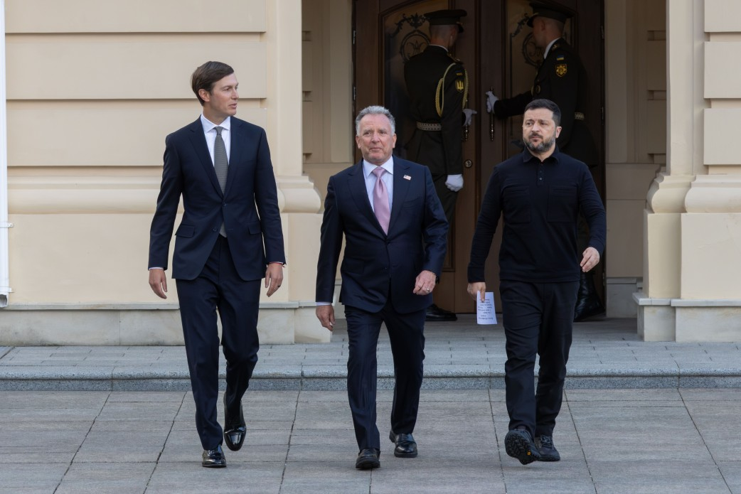 Президент України Володимир Зеленський разом із закордонними гостями йде по брукованому майданчику біля входу до офіційної будівлі у сонячний день. З лівого боку кадру йде Джаред Кушнер у класичному темно-синьому костюмі, білій сорочці, сірій краватці та туфлях. По центру крокує Стів Віткофф, також одягнений у темно-синій костюм, білу сорочку з рожевою краваткою та значком-прапором на лацкані піджака. Праворуч йде Президент України Володимир Зеленський у фірмовій чорній тактичній сорочці-поло з довгими рукавами та чорних штанях, тримаючи в лівій руці невеликий аркуш паперу з нотатками. На задньому плані біля дверей будівлі на ганку стоять двоє військовослужбовців почесної варти Збройних Сил України у парадній формі. Фасад будівлі виконаний у світло-бежевих тонах із класичними колонами та елементами декору.