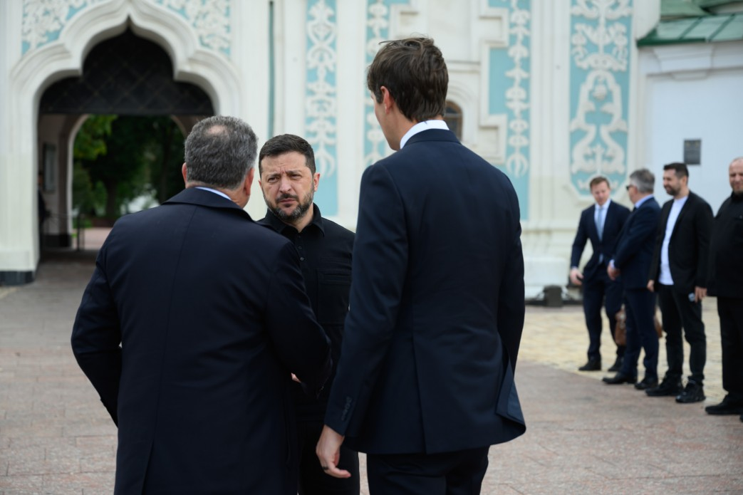 Президент України Володимир Зеленський спілкується зі спецпосланцями США Стівом Віткоффом та Джаредом Кушнером на території історичного комплексу в Києві. Володимир Зеленський, одягнений у чорну тактичну форму, стоїть по центру обличчям до камери й зосереджено слухає співрозмовників. Перед ним спиною до камери стоять Стів Віткофф у темно-синьому пальті та Джаред Кушнер у класичному темному костюмі та білій сорочці. Зустріч проходить на брукованому майданчику просто неба. На задньому плані ліворуч видно старовинну білу аркову браму з блакитним художнім орнаментом, а праворуч у ряд стоять чоловіки з супроводу та охорони в офіційних костюмах.