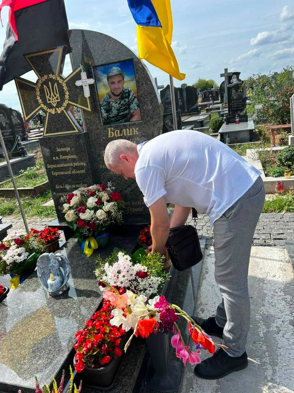Представник Голопристанської громади Херсонської області поправляє живі квіти на могилі загиблого військовослужбовця під час покладання квітів делегаціями до могил воїнів ЗСУ в межах відзначення Дня міста Чорткова. Чоловік у білій футболці та сірих штанях із чорною сумкою через плече схилився з правого боку кадру над могильними плитами у сонячний день на кладовищі. Поховання облаштоване великим сірим гранітним пам'ятником із кольоровим фотопортретом українського захисника у камуфляжній формі та написом Балик. Ліворуч на стелі викарбувано великий козацький хрест із золотим тризубом, під яким нанесено текст про те, що воїн загинув біля населеного пункту Петрівка Бериславського району Херсонської області. На могилі розставлено багато свіжих букетів білих, червоних і рожевих квітів, кошик із синьо-жовтою стрічкою та маленьку фігурку янгола зі свічкою, а зверху над пам'ятником на флагштоках майорять великий державний синьо-жовтий прапор України та червоно-чорний прапор. На задньому плані видно інші надгробки цвинтаря під хмарним небом.