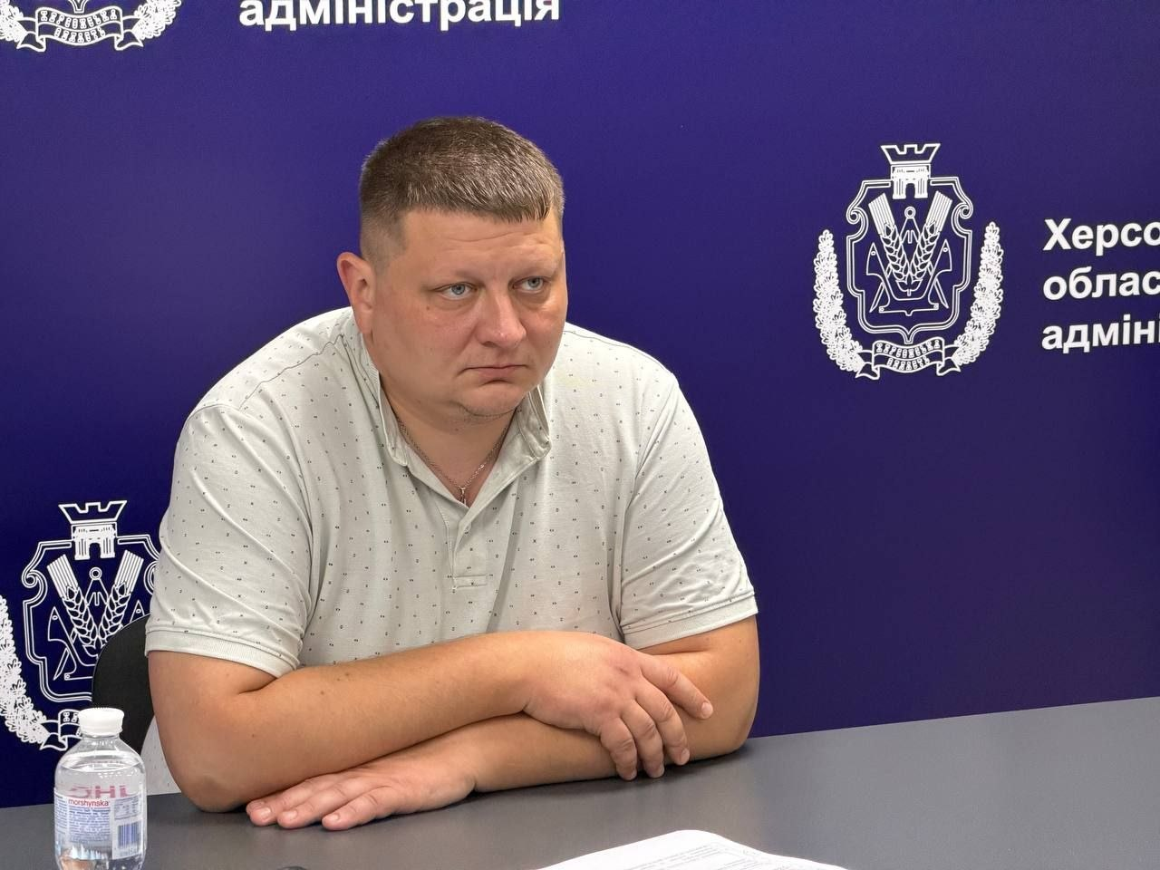 Начальник управління інформаційних технологій Херсонської обласної військової адміністрації Олександр Касіч сидить за сірим столом під час навчально-методичних зборів, присвячених захисту інформаційних систем та зміцненню кібербезпеки в громадах області. Посадовець одягнений у світло-сіру літню сорочку-поло у дрібну цяточку, його руки складені на столі перед собою, а погляд зосереджений під час обговорення критеріїв оцінки стану захищеності державних інформаційних ресурсів. Засідання проходить у службовому приміщенні на тлі офіційного темно-синього банера з повторюваними великими білими емблемами та написами Херсонська обласна державна адміністрація. Навчання організовано для підвищення стійкості цифрової інфраструктури органів місцевого самоврядування до зовнішніх загроз за участю представників Держспецзв’язку та профільних науковців.