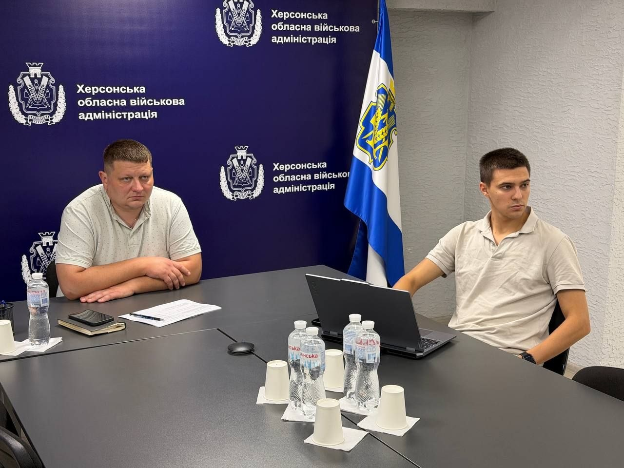 Начальник управління інформаційних технологій Херсонської ОВА Олександр Касіч та його колега — працівник цього ж управління — сидять за великим сірим столом під час навчально-методичних зборів із кібербезпеки та захисту інформаційних систем громад. Ліворуч Олександр Касіч у світло-сірій сорочці-поло у дрібну цяточку сидить, склавши руки перед собою, а перед ним на столі розкладено робочі документи, лежить закритий блокнот, смартфон та стоїть пластикова пляшка мінеральної води. Праворуч за столом сидить молодий співробітник управління у бежевій сорочці-поло, який уважно слухає доповідача перед відкритим чорним ноутбуком. На передньому плані по центру столу розставлено пляшки з водою Моршинська та білі паперові стаканчики. Засідання проходить у кабінеті на тлі офіційного темно-синього банера з написами Херсонська обласна військова адміністрація, а поруч встановлено синьо-білий прапор Херсонської області з офіційним гербом.