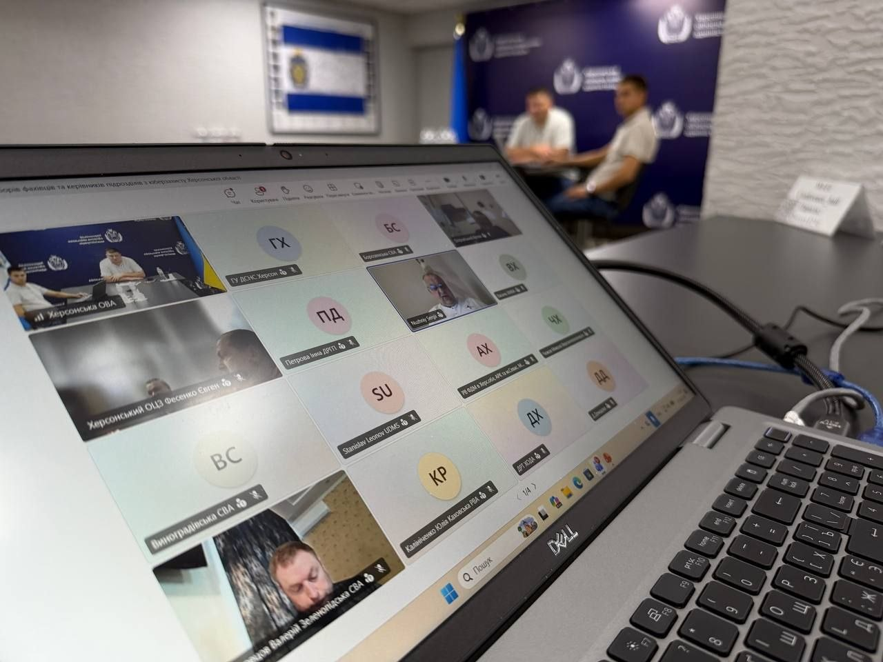 Екран відкритого сріблястого ноутбука Dell, на якому відображається інтерфейс онлайн-конференції під час проведення навчально-методичних зборів для фахівців і керівників підрозділів кіберзахисту громад Херсонщини. На дисплеї комп'ютера чітко видно вікна з відеотрансляціями та текстовими ініціалами підключених учасників наради, серед яких підписи представників органів місцевого самоврядування та державних служб: Херсонська ОВА, ГУ ДСНС Херсонської області, Виноградівська СВА, Зеленопідська СВА, Каланчацька СВА та інші територіальні підрозділи. Ноутбук стоїть на чорному офісному столі, а з правого боку до нього підключено робочий кабель. На задньому плані кадру з розмиттям фокусу видно робочий кабінет, де за великим столом на тлі темно-синього брендованого банера Херсонської ОВА та синьо-білого прапора області сидять начальник управління інформаційних технологій Олександр Касіч та його співробітник, які організували цей практичний інтенсив для підвищення стійкості цифрової інфраструктури до зовнішніх загроз.