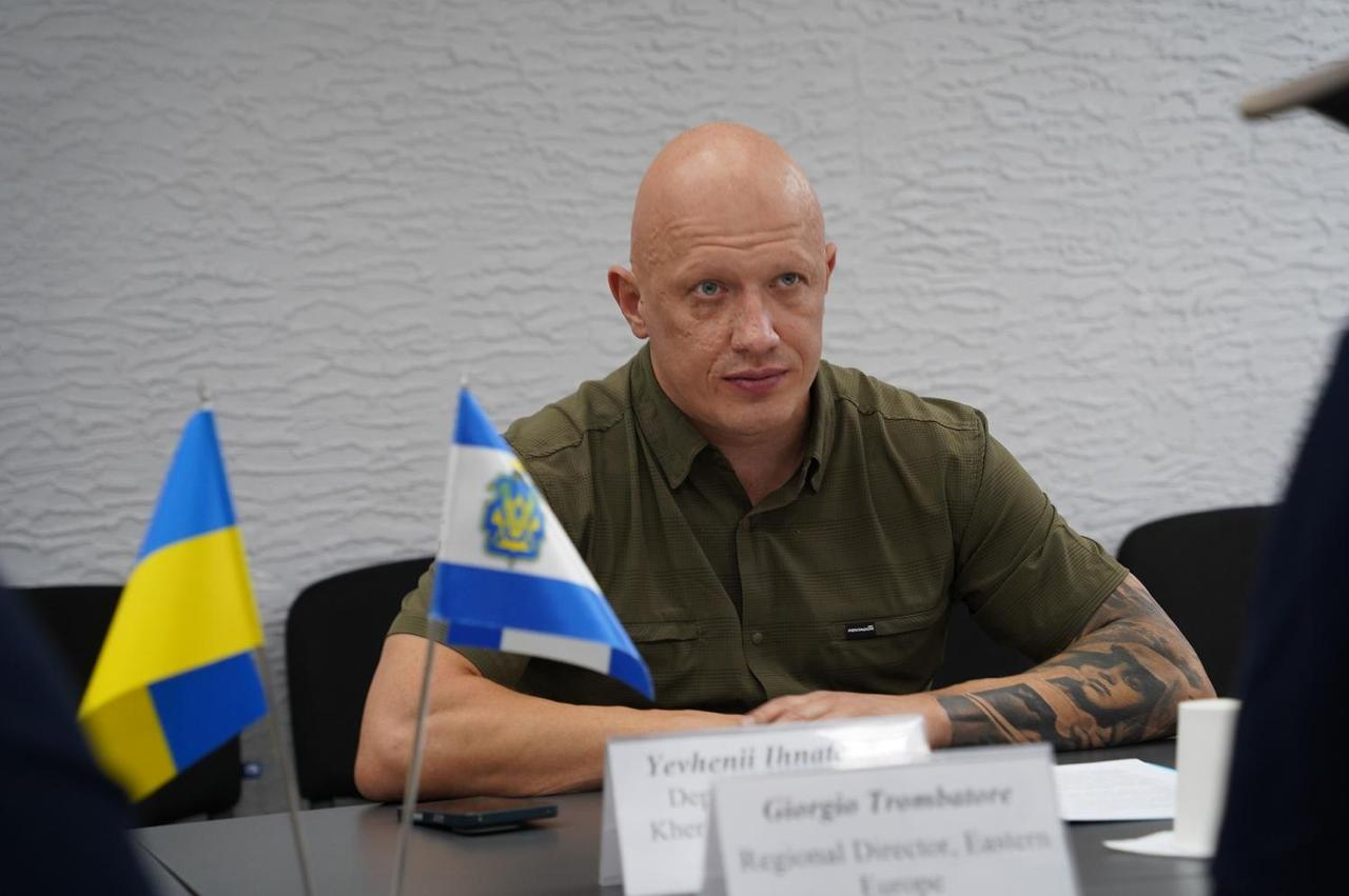 Заступник начальника Херсонської обласної військової адміністрації Євгеній Ігнатенко сидить за великим робочим столом під час офіційної зустрічі з делегацією міжнародної неурядової організації Project HOPE, де обговорювали підтримку та розвиток медичної галузі області в умовах війни. Посадовець одягдений у тактичну сорочку кольору хакі з коротким рукавом. Він зосереджено дивиться перед собою, слухаючи пропозиції закордонних партнерів щодо оновлення лікувально-діагностичного обладнання та забезпечення автономного водопостачання в медзакладах. На столі ліворуч встановлено два настільні прапорці — державний синьо-жовтий прапор України та біло-синій прапор Херсонської області з офіційним гербом, а засідання проходить на тлі світло-сірої фактурної стіни кабінету.