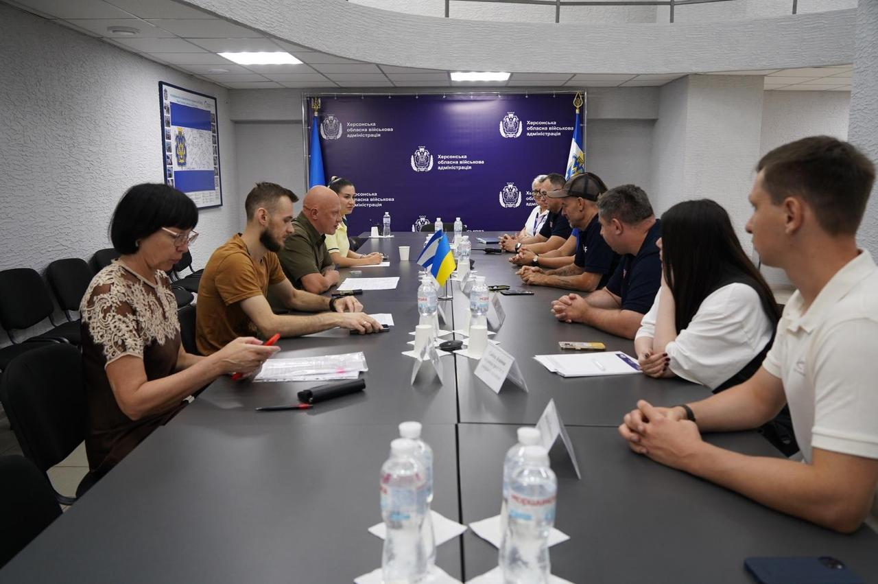 Офіційне засідання та двосторонні переговори у просторій залі Херсонської обласної військової адміністрації, присвячені обговоренню підтримки медичної галузі області, оновленню лікувально-діагностичного обладнання та бурінню свердловин для автономного водопостачання лікарень в умовах війни. Понад десять учасників зустрічі сидять один навпроти одного по обидва боки довгого темного столу. З лівого боку столу розмістилися українські посадовці. Першою ліворуч на передньому плані сидить заступниця Херсонського міського голови Алла Малицька у коричневій блузі з білим мереживом, поруч із нею — заступник директора Департаменту здоров’я Михайло Балацький у коричневій футболці, далі — заступник начальника Херсонської ОВА Євгеній Ігнатенко у тактичній сорочці кольору хакі, а на дальньому плані за ними сидить начальниця управління міжнародного співробітництва та залучення інвестицій Херсонської ОДА Марія Богодухова у світлій сорочці. З правого боку столу сидять члени делегації міжнародної неурядової організації Project HOPE у фірмових темно-синіх сорочках-поло, а на передньому плані праворуч — представниця організації у білій блузі та її колега - молодий чоловік у світлій футболці-поло. На столі перед присутніми розставлено пластикові пляшки з мінеральною водою, паперові стаканчики, розкладено робочі папери, документи, іменні таблички, а по центру встановлено маленькі настільні прапорці України та Херсонської області. Засідання проходить на тлі великого темно-синього офіційного банера з повторюваними гербами та білими написами Херсонська обласна військова адміністрація.