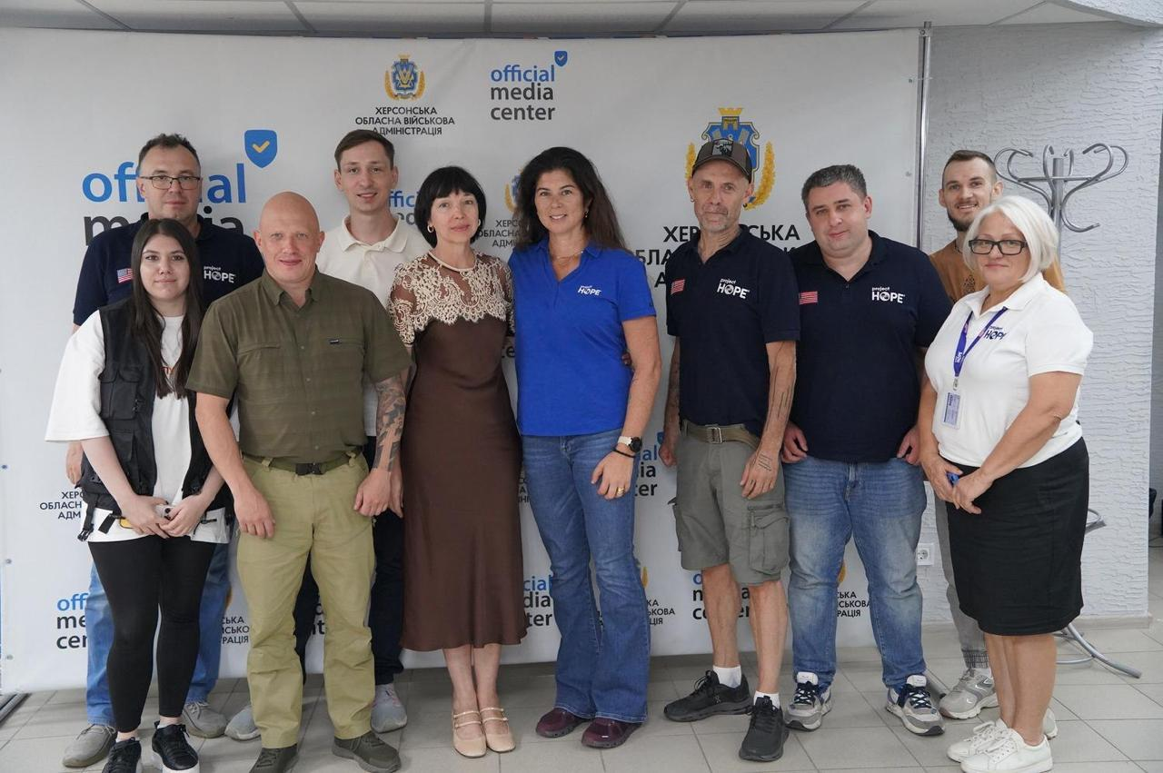 Групова фотографія одинадцяти учасників офіційної зустрічі між керівництвом Херсонщини та делегацією міжнародної неурядової організації Project HOPE, присвяченої підтримці медичної галузі області, оновленню обладнання та бурінню свердловин для автономного водопостачання лікарень. Чоловіки та жінки стоять у ряд на тлі великого білого банера з написом Херсонська обласна військова адміністрація та синіми логотипами Офіційний media center англійською мовою. Третім ліворуч у першому ряду стоїть заступник начальника Херсонської ОВА Євгеній Ігнатенко у тактичній сорочці та штанях кольору хакі. Поруч із ним стоїть заступниця Херсонського міського голови Алла Малицька у довгій коричневій сукні з білим мереживом. По центру кадру стоїть віцепрезидентка з глобальних програм та операцій організації Project HOPE Пія Ванек у синій фірмовій футболці-поло та джинсах. Праворуч від неї стоять інші члени закордонної делегації у темно-синіх сорочках-поло з білими логотипами проєкту, зокрема регіональний директор Джорджіо Тромбаторе у кепці та сірих шортах, а біля нього — представниця організації в окулярах та білій футболці. Крайній праворуч у другому ряду, за спинами колег, стоїть заступник директора Департаменту здоров’я Михайло Балацький у коричневій футболці.