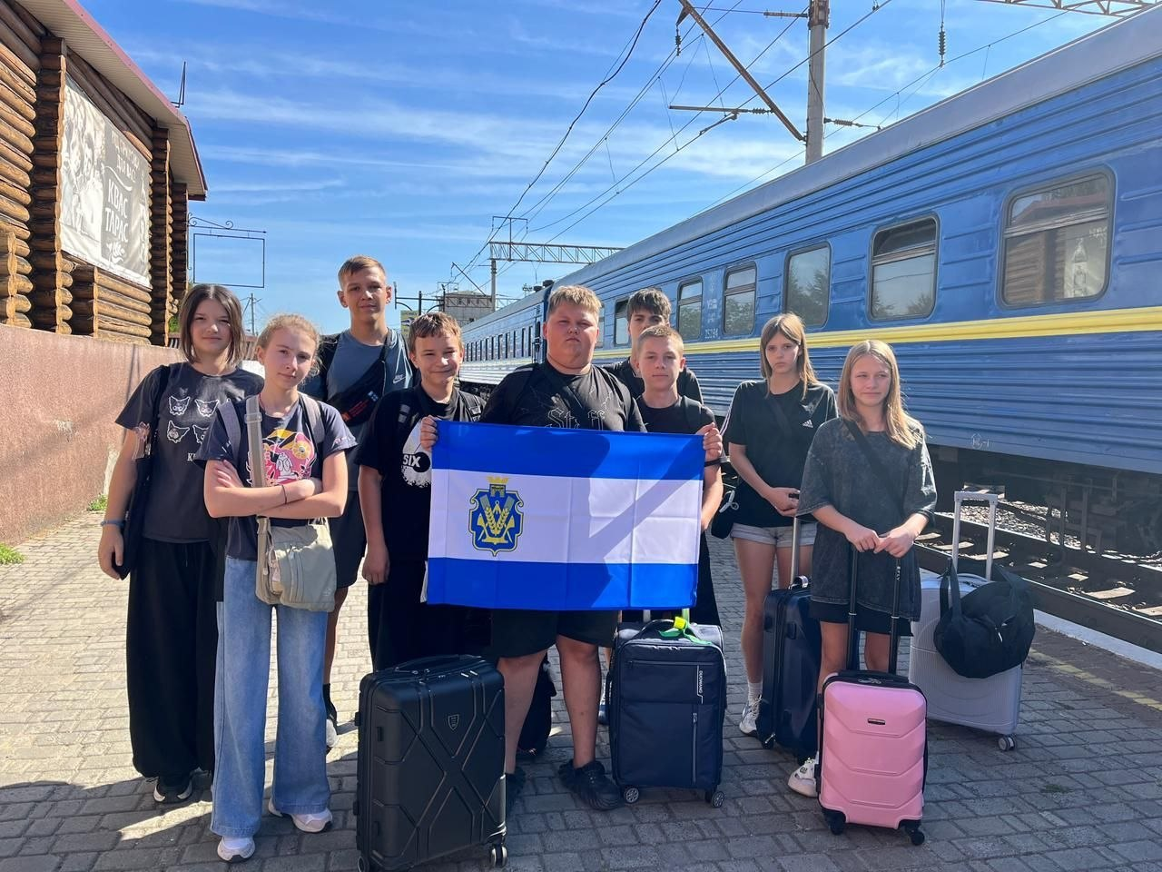 Група підлітків із Херсонщини з дорожніми валізами позує на залізничному пероні перед відправленням до літнього табору в Угорщині. Діти в літньому одязі стоять на тлі синього пасажирського вагона Укрзалізниці. Хлопці по центру тримають розгорнутий синьо-біло-синій прапор Херсонської області з гербом у лівій частині. Юні українці вирушають на відпочинок, екскурсії та психологічне розвантаження до міста Шаторальяуйхей за підтримки Асоціації імені Ракоці та Генерального консульства Угорщини в Ужгороді. Програма відпочинку включає відвідування замку Фюзер, прогулянки Мостом Національної єдності, заняття з ремесел, спортивні та інтелектуальні заходи для дітей, які постраждали внаслідок війни. На задньому плані фото видно залізничні електромережі, дерев'яну будівлю та чисте блакитне небо.