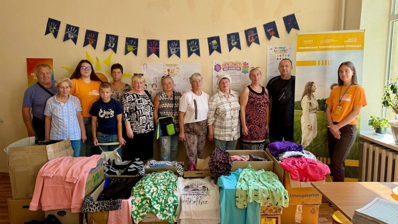 Внутрішньо переміщені особи з Херсонщини позують для спільного фото у світлому кабінеті кропивницького координаційного центру Вільні разом під час отримання допомоги від благодійної організації Asociación Ucrania - Rioja. Переселенці різного віку стоять у ряд, радо посміхаючись кадру. На передньому плані перед ними розставлено великі картонні коробки, які вщерть заповнені новим дитячим та підлітковим одягом, де видно рожеву кофту, зелену піжаму, спортивні костюми, футболки та штани. Крайня праворуч стоїть молода волонтерка центру в офіційній помаранчевій футболці та темних штанях із бейджем на шиї. На задньому плані на світлій стіні висить паперова гірлянда з синіх прапорців із малюнками долоньок, дитячі інформаційні плакати, малюнок жовтого сонця та кольоровий чек-лист. Праворуч у кутку встановлено великий інформаційний ролап-стенд Рівненської територіальної громади жовто-білого кольору, а біля вікна праворуч на підвіконні стоять зелені кімнатні рослини в горщиках.