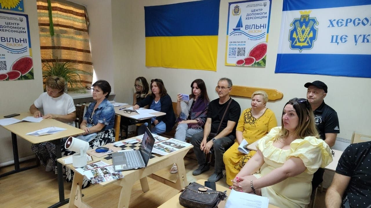 Внутрішньо переміщені особи з Херсонщини сидять на стільцях та за партами у світлому кабінеті кропивницького координаційного центру Вільні разом під час навчання з мінної безпеки, організованого спільно з фахівцями Товариства Червоного Хреста України. Переселенці зосереджено слухають лектора та дивляться вперед, вивчаючи правила поводження з вибухонебезпечними предметами. На передньому плані по центру стоїть невеликий дерев'яний столик, на якому розкладено друковані інформаційні брошури та пам'ятки, встановлено білий мультимедійний проєктор та відкритий сріблястий ноутбук. Ліворуч за партами сидять жінки, одна з яких робить нотатки на папері. На задньому плані кадру на світлій стіні розвішано офіційну символіку: великий державний прапор України, прапор Херсонської області з офіційним регіональним гербом та великим білим написом Херсон це Україна, а також інформаційні плакати з логотипом Центр допомоги херсонцям Вільні разом та малюнком кавуна.