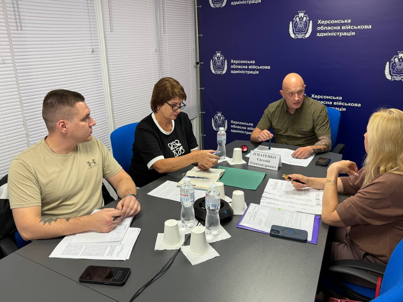 Заступник начальника Херсонської ОВА Євгеній Ігнатенко, заступниця начальника Херсонської МВА Наталія Чорненька та фахівці профільних структурних підрозділів під час онлайн-підключення до урядової наради Міністерства соціальної політики щодо підтримки населення деокупованих громад під час опалювального сезону. Четверо учасників зустрічі від області сидять за кутовим темним столом у кабінеті, обговорюючи поточну ситуацію із забезпеченням жителів паливом та виплатами. На дальньому плані по центру сидить Євгеній Ігнатенко у тактичній сорочці кольору хакі. Ліворуч за столом розмістилися двоє колег: молодий співробітник ОВА у бежевій футболці з документами в руках та Наталія Чорненька в окулярах і чорній кофті з білим орнаментом, яка робить нотатки у відкритому блокноті. Праворуч на передньому плані спиною до кадру сидить фахівчиня у коричневій кофті з довгим світлим волоссям, яка вивчає розкладені папери з даними моніторингу потреб осель. Нарада проходить на тлі великого темно-синього офіційного банера з написом Херсонська обласна військова адміністрація, а ліворуч видно закриті білі віконні жалюзі.