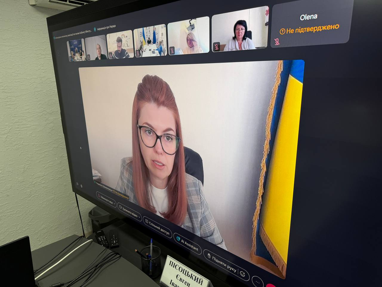 Екран великого чорного монітора комп'ютера, на якому відображається інтерфейс онлайн-відеоконференції під час урядової наради Міністерства соціальної політики щодо забезпечення паливом та виплатами жителів деокупованих громад Херсонщини на прийдешню зиму. На дисплеї крупним планом показано вікно головної доповідачки — представниці міністерства в окулярах із темною оправою та в сірому піджаку в клітинку, яка виступає на тлі державного прапора України із золотистою бахромою. У верхній частині екрана розташовано горизонтальний ряд із меншими віконцями інших підключених учасників, серед яких кабінети регіональних посадовців та заставка з іменем Olena. Перед монітором на темному робочому столі в кабінеті встановлено іменну табличку, на якій видно напис Пісоцький Євген Іванович. Вона належить директору Департаменту з питань цивільного захисту та оборонної роботи Херсонської ОДА, який долучився до обговорення потреб тридцяти шести з половиною тисяч осель області.