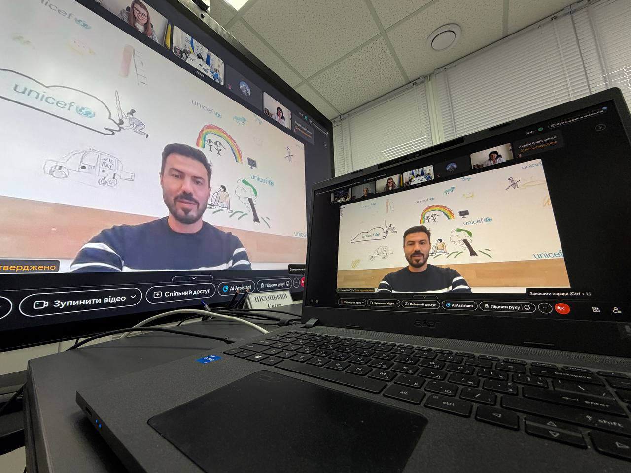 Екран відкритого чорного ноутбука Acer та великого настільного монітора комп'ютера, на яких дублюється інтерфейс онлайн-відеоконференції під час урядової наради Міністерства соціальної політики щодо забезпечення паливом та виплатами жителів деокупованих громад Херсонщини на прийдешню зиму. На обох дисплеях крупним планом показано вікно виступу одного з учасників онлайн-підключення — темнобрового чоловіка з короткою бородою у темно-синьому світшоті з горизонтальними білими смугами на рукавах. Він виступає на тлі світлої стіни з дитячими малюнками та логотипами міжнародної організації ЮНІСЕФ, яка також долучилася до програм підтримки родин. На столі між ноутбуком та монітором у кабінеті встановлено іменну табличку, на якій видно напис Пісоцький Євген Іванович. Вона належить директору Департаменту з питань цивільного захисту та оборонної роботи Херсонської ОВА, який бере участь в обговоренні потреб тридцяти шести з половиною тисяч осель деокупованих територій. 