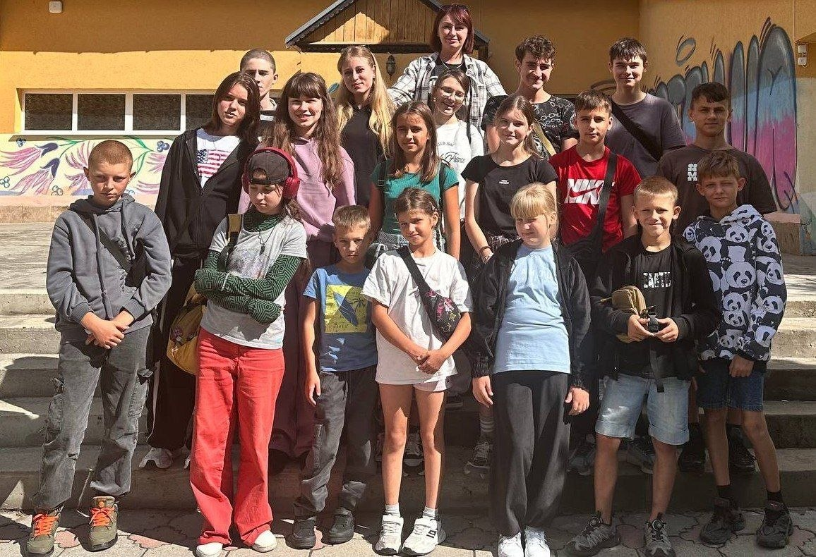 Група дітей та підлітків із різних громад Херсонщини позує для спільного фото під час відправлення на оздоровлення та відпочинок до Міжнародного дитячого центру Артек, що розташований у селі Березники на Закарпатті. Діти зі статусом ВПО, які наразі проживають у різних областях України, стоять разом у літньому одязі на сходах будівлі в сонячний день. Поїздку організовано за сприяння Департаменту соціального розвитку Херсонської ОДА в межах дев’ятої тематичної зміни, присвяченої профорієнтаційному самовизначенню. Програма відпочинку тривалістю у 21 день спрямована на психологічне розвантаження, отримання яскравих вражень та соціалізацію дітей, які постраждали внаслідок війни.