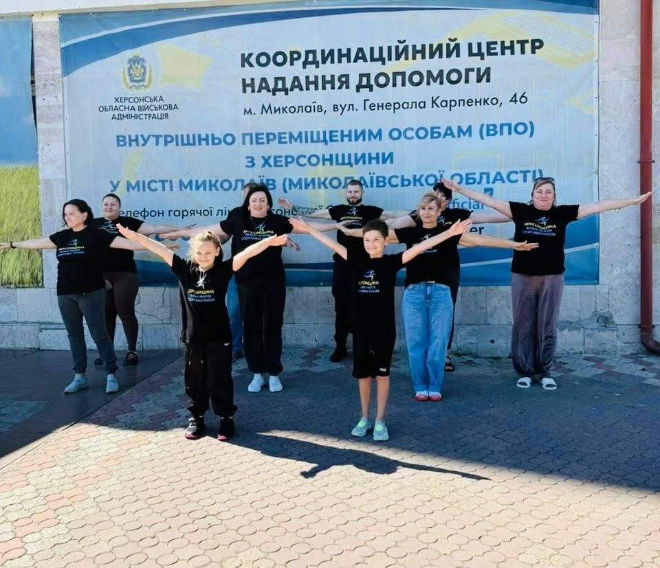 Група дорослих та дітей у чорних брендованих футболках робить фізичну руханку на свіжому повітрі перед входом до будівлі, розвівши руки в сторони. Вони позують на тлі великого інформаційного банера Херсонської обласної військової адміністрації з написом Координаційний центр надання допомоги внутрішньо переміщеним особам (ВПО) з Херсонщини у місті Миколаїв. Захід присвячений старту Тижня спорту в координаційному центрі Вільні разом, до якого долучилися фахівці та відвідувачі простору. Програма спрямована на підтримку здорового способу життя, зміцнення фізичного й ментального здоров'я та психологічне розвантаження переселенців, і включає руханки, конкурси малюнків на дитячому артмайданчику, книжковий куточок та інформаційні години про історію українського спорту.