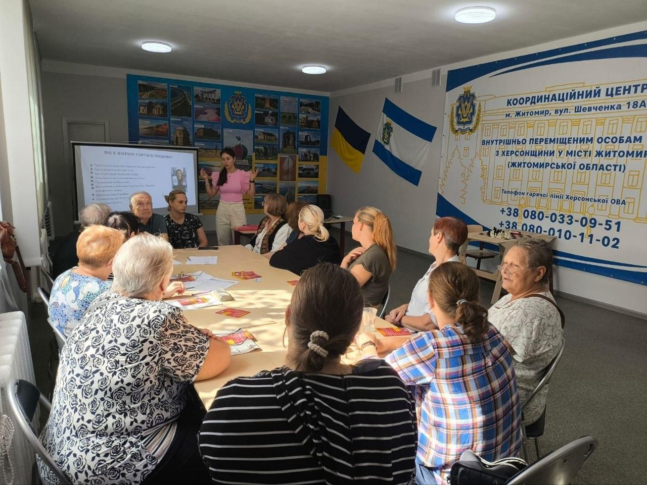 Група людей старшого віку та дорослих сидить за великим столом у залі хабу та слухає лекторку під час інформаційного заходу щодо психологічної та соціальної допомоги. Дівчина в рожевій кофті стоїть біля інтерактивної дошки та розповідає матеріал, показуючи слайд із переліком правил безпеки. Праворуч на стіні розміщено великий інформаційний банер із написом Координаційний центр внутрішньо переміщеним особам з Херсонщини у місті Житомир (Житомирської області), а також номери телефонів гарячої лінії Херсонської ОВА. Поруч на стіні висять державний прапор України та прапор Херсонської області. Захід є частиною системної підтримки переселенців у координаційному центрі Вільні разом, де представниця громадської організації Авенір провела тренінг-лекцію з протидії торгівлі людьми та безпечного працевлаштування з метою захисту прав громадян, надання психологічної допомоги та підвищення життєстійкості внутрішньо переміщених осіб.