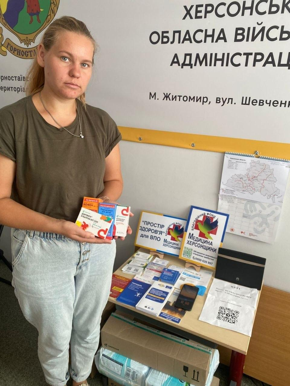 Молода жінка стоїть у приміщенні хабу та тримає в руках отримані пакування з ліками, серед яких дитячий сироп Бофен та інші медичні препарати. Вона позує поруч зі столом, на якому розміщено буклет із написом Простір здоров'я для ВПО Медицина Херсонщини та інформаційні картки благодійного фонду Медицина Херсонщини із зображенням кольорового птаха. На стіні позаду жінки видно частину великого білого банера Херсонської обласної військової адміністрації з написом м. Житомир, а також герб Горностаївської селищної територіальної громади та карту. Захід є частиною системної підтримки переселенців у координаційному центрі Вільні разом у Житомирі, де для отримання допомоги зареєстрували 11 переселенців. Видача ліків та засобів гігієни від благодійного фонду Медицина Херсонщини спрямована на підтримку здоров'я, покращення соціально-побутового благополуччя та підвищення життєстійкості внутрішньо переміщених осіб із Херсонської області.