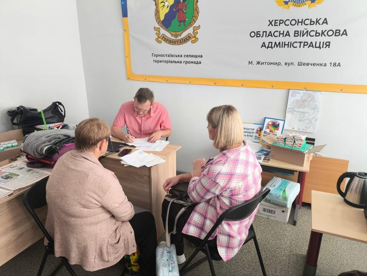 Чоловік у рожевій сорочці сидить за робочим столом у кабінеті хабу та заповнює документи, надаючи правову допомогу двом жінкам, які сидять навпроти нього на стільцях. На столі розкладені папери та мобільний телефон, а поруч на полицях видно коробки з медикаментами, засобами гігієни та буклет Простір здоров'я для ВПО Медицина Херсонщини. На стіні позаду спеціаліста розміщено великий білий банер із написом Херсонська обласна військова адміністрація, м. Житомир, а також офіційний герб із написом Горностаївська селищна територіальна громада. Захід є частиною юридичної допомоги, яку системно надає координаційний центр Вільні разом у Житомирі для підтримки переселенців із Херсонщини. Індивідуальні консультації, які надав юрисконсульт благодійної організації Благодійний фонд Право на захист для 5 осіб, спрямовані на правовий захист громадян, вирішення юридичних питань та підвищення життєстійкості внутрішньо переміщених осіб.
