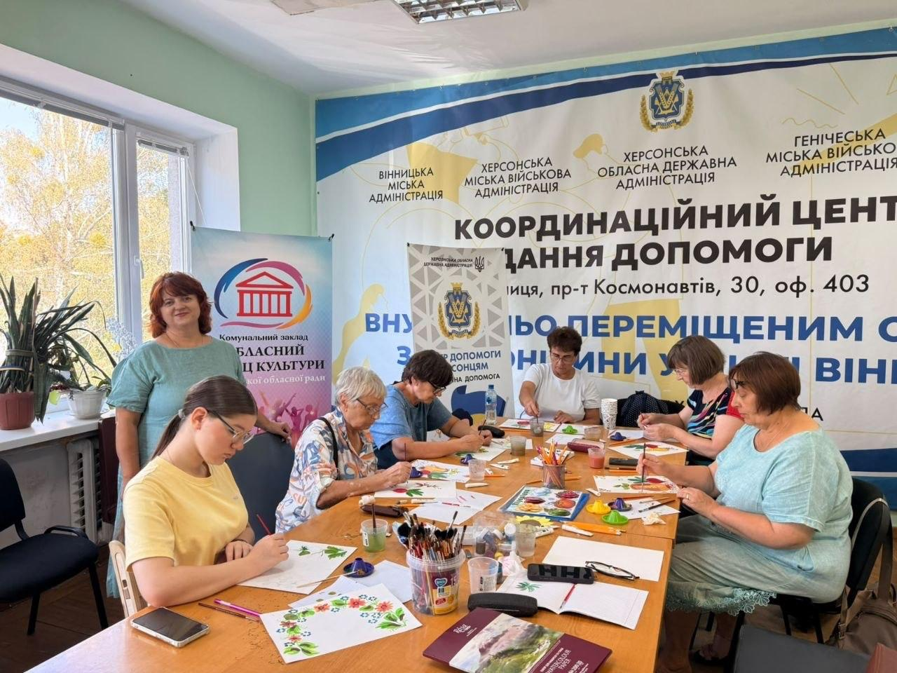 Група жінок та дівчат сидить навколо великого столу в залі хабу та займається творчістю під час майстер-класу з малювання. Учасниці різного віку акуратно виводять пензликами візерунки з квітів, ягід та калини на білих аркушах паперу, використовуючи гуаш та акварельні палітри. Поруч стоїть жінка-наставниця в зеленій блузі, яка спостерігає за процесом. На задньому плані видно великий банер Координаційного центру надання допомоги внутрішньо переміщеним особам з Херсонщини у місті Вінниця, а також ролап Обласного палацу культури Херсонської обласної ради. Захід є частиною організації дозвілля в координаційному центрі Вільні разом, де відбулось заняття із малювання з елементами традиційного петриківського розпису Казкові овочі з метою арттерапії, збереження культурної спадщини, підтримки ментального здоров'я та психологічного розвантаження внутрішньо переміщених осіб.