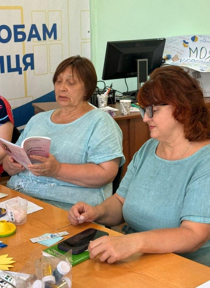 Дві жінки сидять поруч за робочим столом у приміщенні хабу під час проведення культурного заходу. Жінка ліворуч у блакитній футболці зосереджено читає вголос тексти з невеликої книги у рожевій обкладинці, тримаючи її в руках, а її сусідка в окулярах та зеленій блузі уважно слухає читання. На столі перед ними розкладені матеріали для творчості, баночки з блискітками та мобільний телефон, а на задньому плані видно офісний стіл із комп'ютерним монітором та великий білий банер Координаційного центру надання допомоги внутрішньо переміщеним особам з Херсонщини. Захід організовано в межах роботи координаційного центру Вільні разом у Вінниці, де відбулася літературна зустріч Поетична година, присвячена митцям та поетам із Херсонщини, що спрямовано на збереження культурної ідентичності, єднання земляків, надання психологічної підтримки та емоційне розвантаження переселенців.