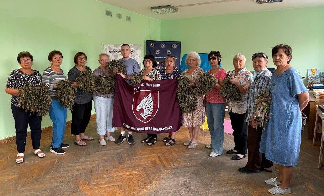 Група майстринь разом із молодим військовослужбовцем позує для спільного фото у світлій залі хабу з паркетною підлогою. Жінки тримають у руках виготовлені власноруч засоби маскування — сплетені з ниток та клаптиків тканини захисні нашоломники. По центру військовий та одна з майстринь розгортають великий бордовий прапор підрозділу із зображенням білого крила на щиті, написом 237 та гаслом Земля для всіх, небо для обраних. На задньому плані видно синій інформаційний банер Проєкт та прапори громад. Патріотична акція відбулася в координаційному центрі Вільні разом у Вінниці, де переселенки з Херсонщини передали представнику 237-ї бригади ЗСУ 22 нашоломники, 5 килимків та 5 поробок-оберегів. Захід ілюструє єдність громади, активну волонтерську підтримку Сил оборони України та залучення внутрішньо переміщених осіб до спільної праці задля наближення перемоги.