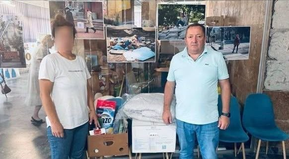 Чоловік та жінка позують у приміщенні хабу на тлі виставки фотографій, які ілюструють наслідки воєнних дій. Праворуч стоїть начальник Чулаківської сільської військової адміністрації Руслан Русаль у світло-блакитній футболці-поло та синіх джинсах. Ліворуч стоїть жінка у білій футболці, яка представляє родину, повернуту з тимчасово окупованих територій (її обличчя розмите з міркувань конфіденційності). Перед ними розкладено гуманітарну допомогу: картонну коробку з великим пакуванням прального порошку та побутовими речами, а також великий пакунок із ковдрою чи постільним набором. Зустріч відбулася в координаційному центрі Вільні разом у Миколаєві в межах програми супроводу та подальшої соціальної підтримки сімей із дітьми, які нещодавно виїхали з громад Херсонщини та знайшли прихисток на Миколаївщині.