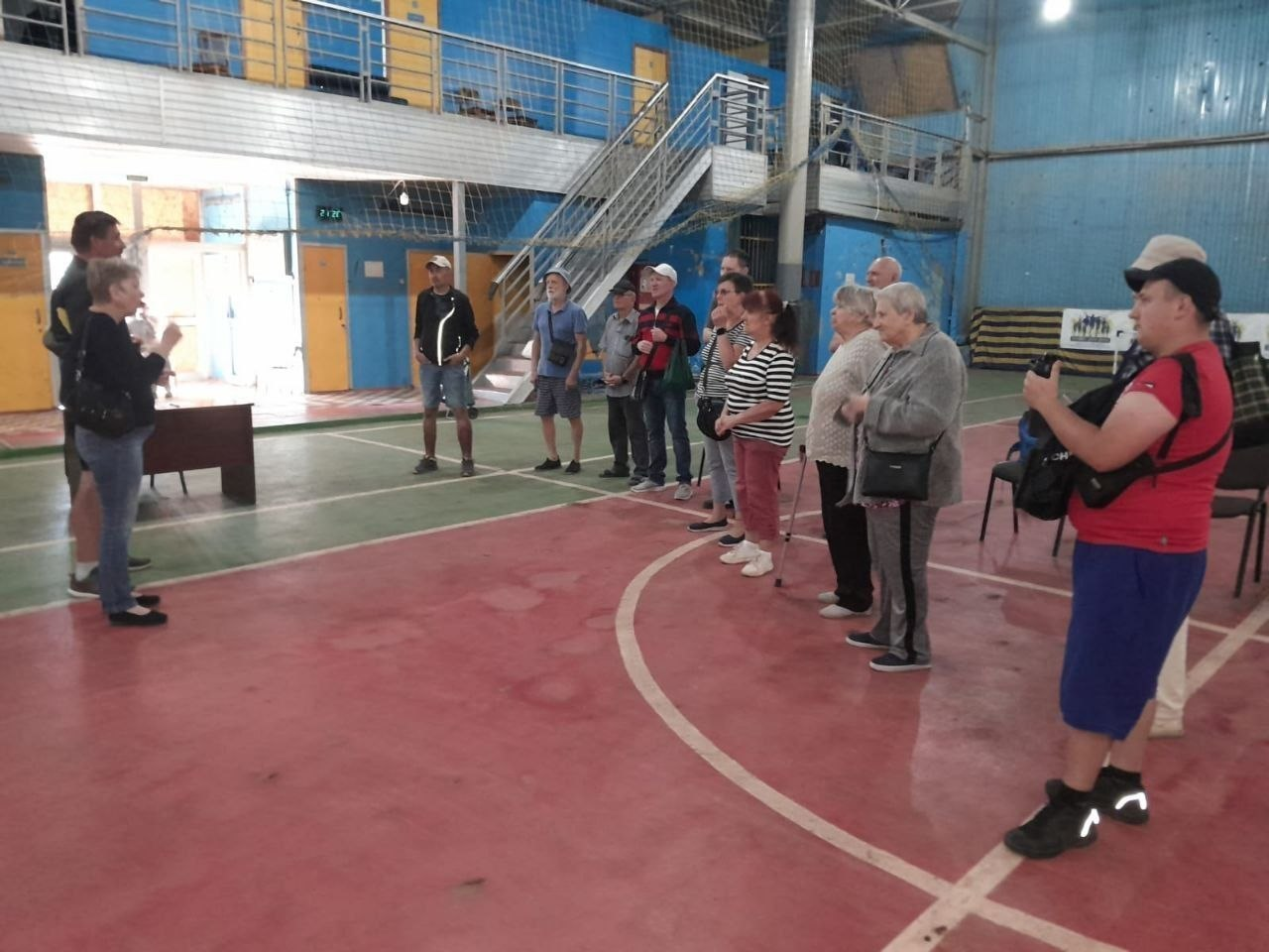 Група людей похилого віку та дорослих слухає інструктаж перед початком змагань у критому спортзалі з червоно-зеленим покриттям підлоги та синіми стінами. Учасники стоять півколом на ігровому майданчику, а ліворуч біля суддівського столу їм пояснюють правила чи програму заходу. На задньому плані видно металеві сходи, що ведуть на балкон другого ярусу будівлі. Фотографія ілюструє організацію змагань із боча серед людей з порушеннями слуху, які відбулися на Херсонщині до Дня фізичної культури і спорту України. Турнір спрямований на залучення людей з інвалідністю до адаптивного спорту, підтримання їхньої фізичної активності, соціалізацію та психоемоційне розвантаження.